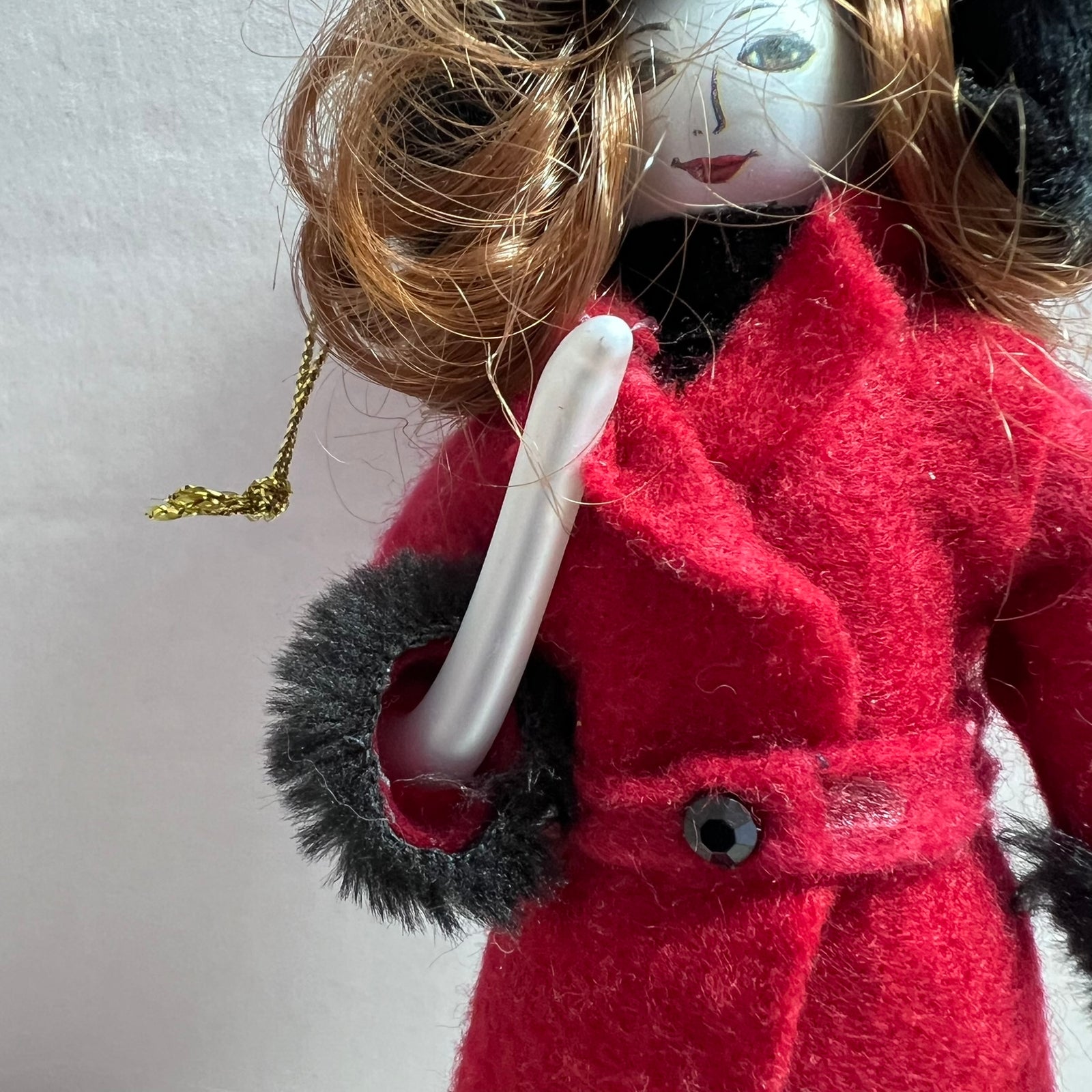 A close-up of a doll wearing a bright red coat with dark gray faux-fur trim, holding a white object, against a plain light background.