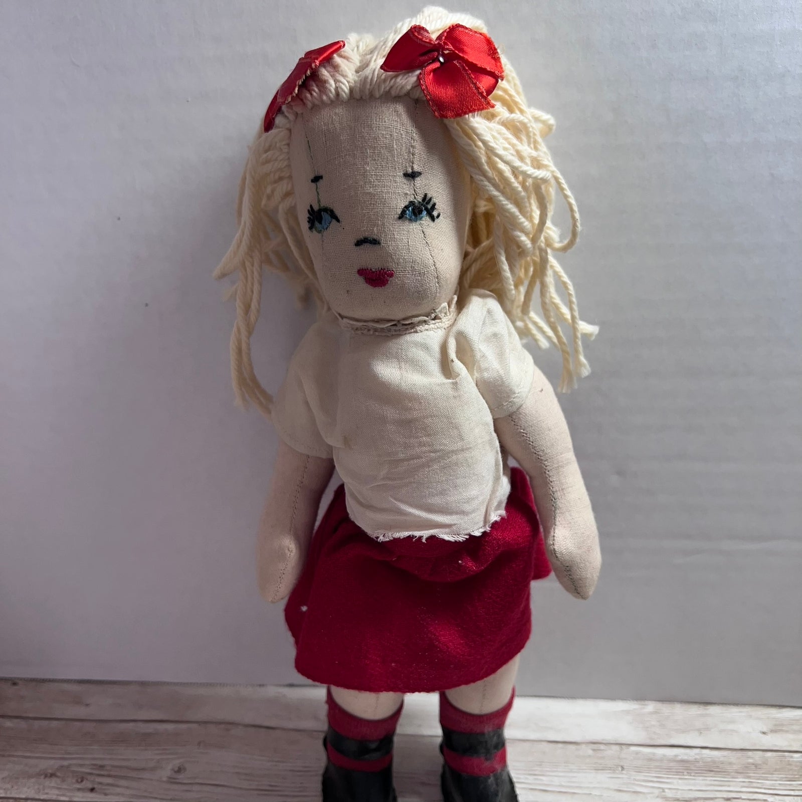 A handmade fabric doll with blonde yarn hair, two red bows, a cream shirt, a red skirt, and striped socks, standing against a plain light gray wall.
