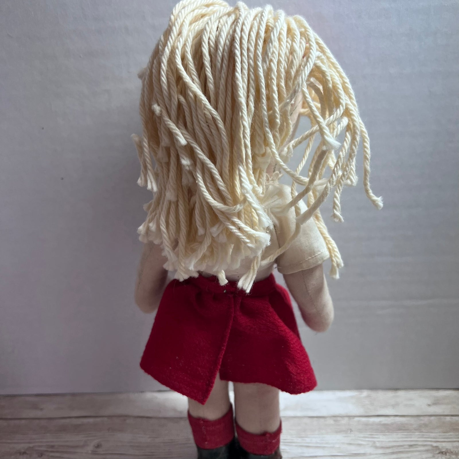 Back view of a handmade doll with messy cream yarn hair and a red skirt, standing on a light wood floor against a gray wall.