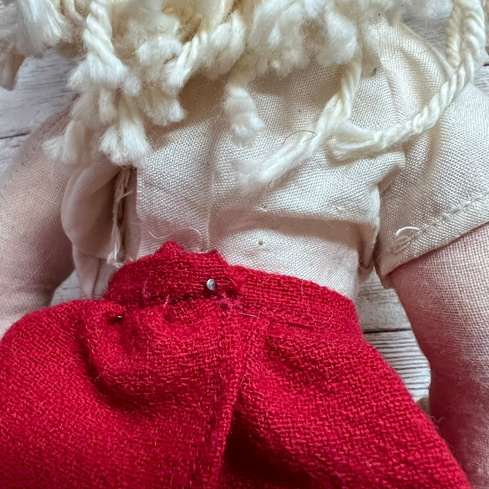 Close-up of a handmade fabric doll or rag doll showing cream-colored linen clothing, a bright red skirt, and textured off-white yarn hair, pinned together on a light wood background.