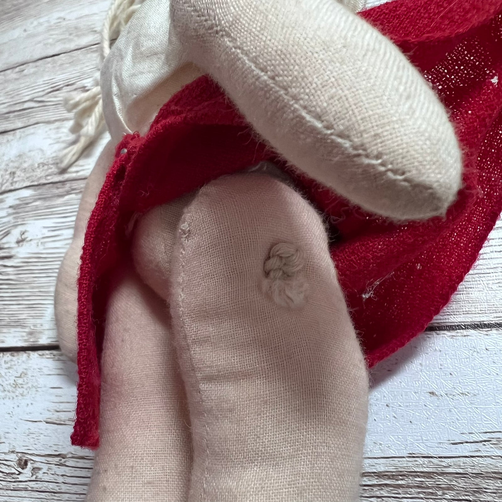 A close-up of a handmade fabric doll or plush toy with cream-colored linen body parts and a red mesh fabric hat or scarf, resting on a white-washed wooden surface.