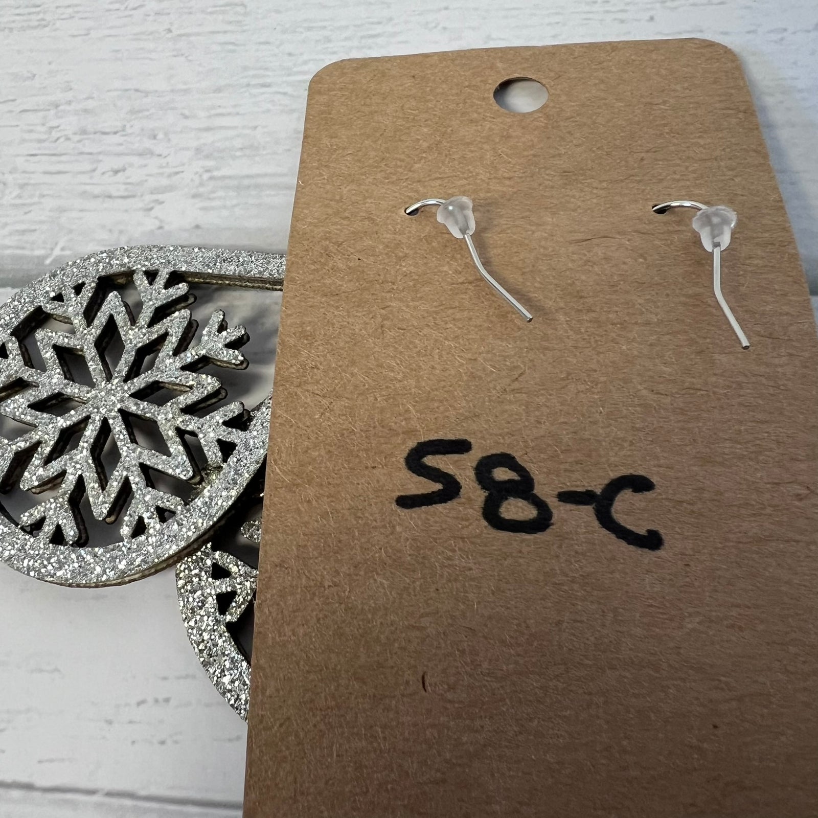 Clear earrings on a brown cardboard tag with 'S8-C' marking, snowflake ornament in the background.
