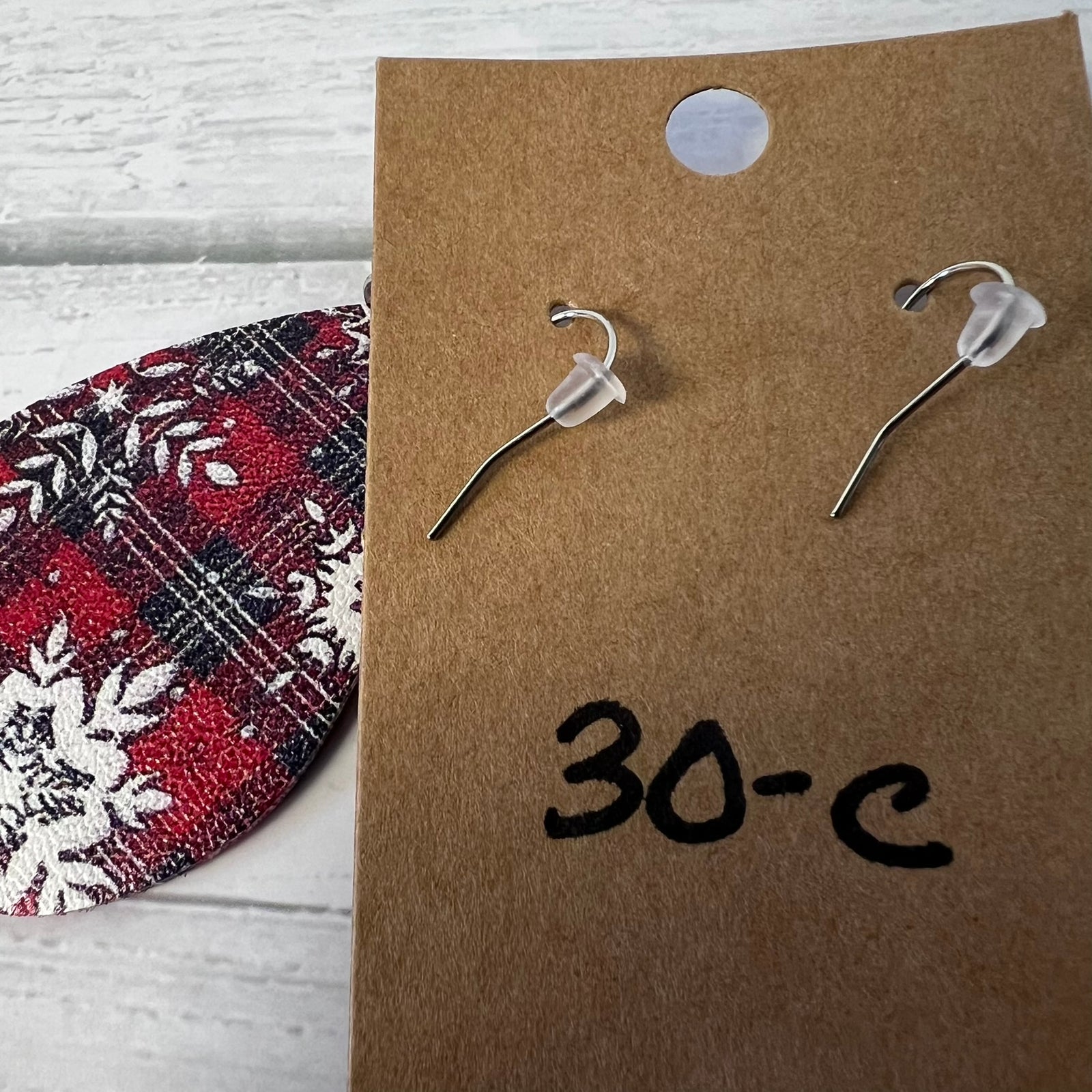 Pair of silver earrings on a brown card with '30-C' written on it, next to a red and white patterned object.