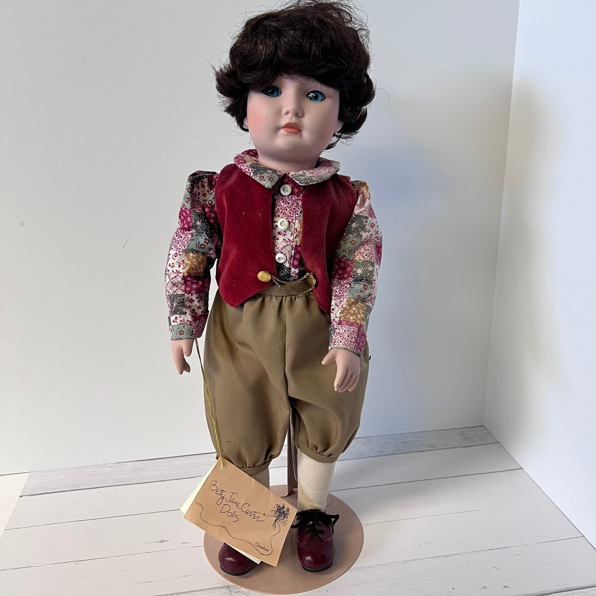 A vintage porcelain doll with dark curly hair, wearing a red vest, patterned shirt, tan trousers, and dark red shoes, standing on a round base with a handwritten tag attached.