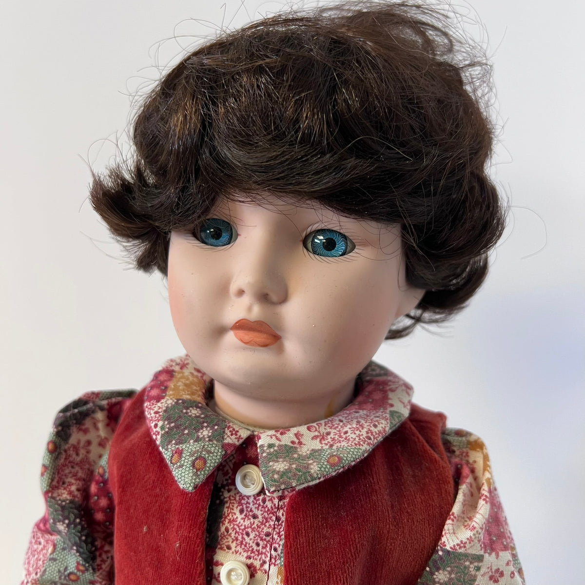 A vintage porcelain doll with dark brown hair, blue eyes, and a red vest over a floral dress, photographed against a plain light background.