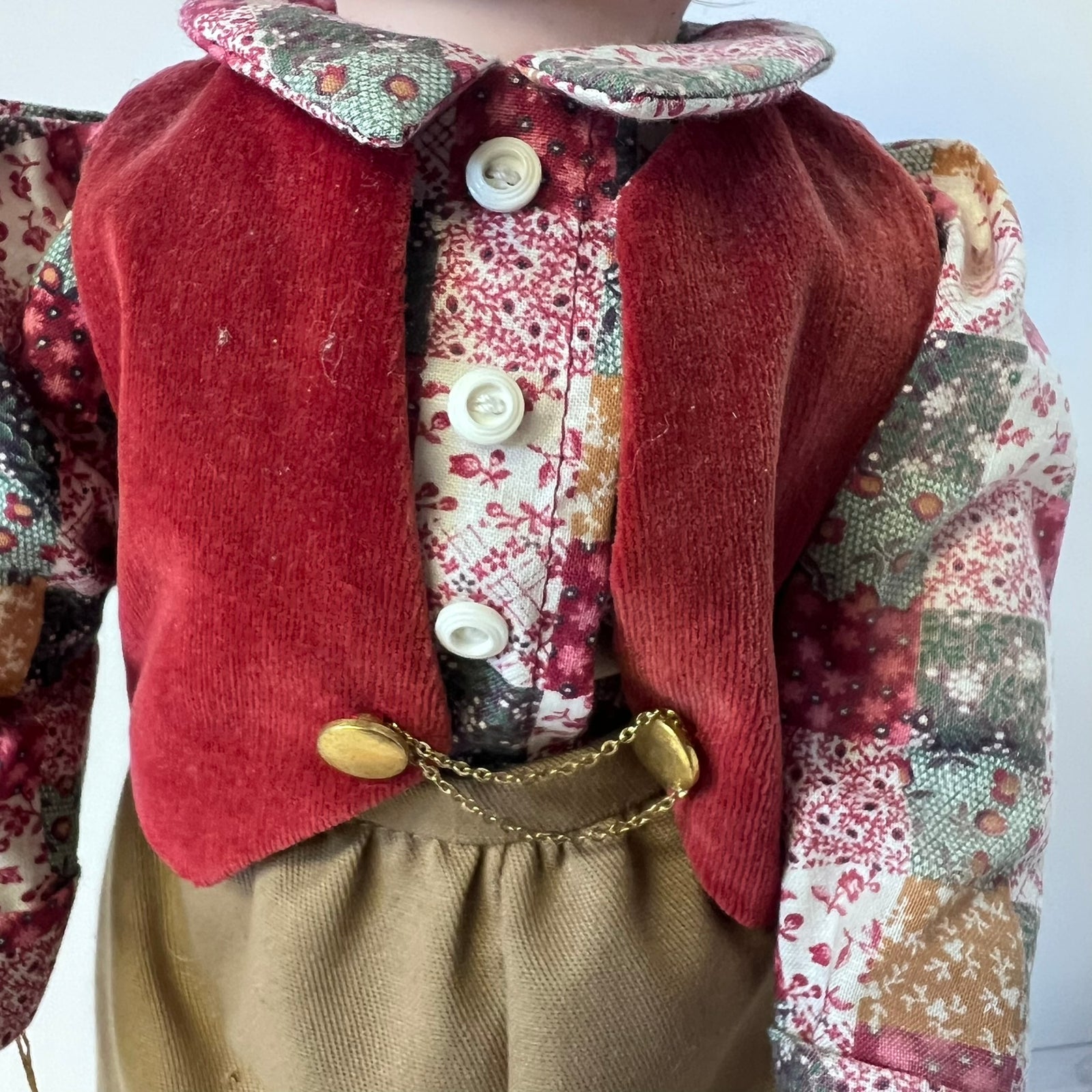 A close-up of a vintage doll or toy dressed in a red corduroy vest over a floral patchwork shirt and tan corduroy trousers, with a gold chain detail.