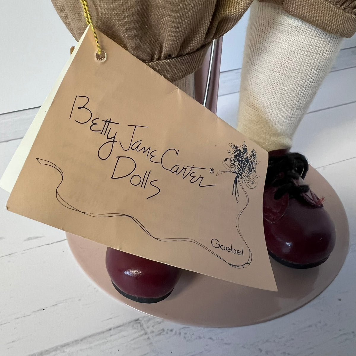 A close-up of a Betty Jane Carter Dolls figure showing the lower half, including a tan skirt, cream fabric legs, dark red shoes, and a hanging paper tag that reads 'Betty Jane Carter Dolls' with the brand 'Goebel' printed on it.