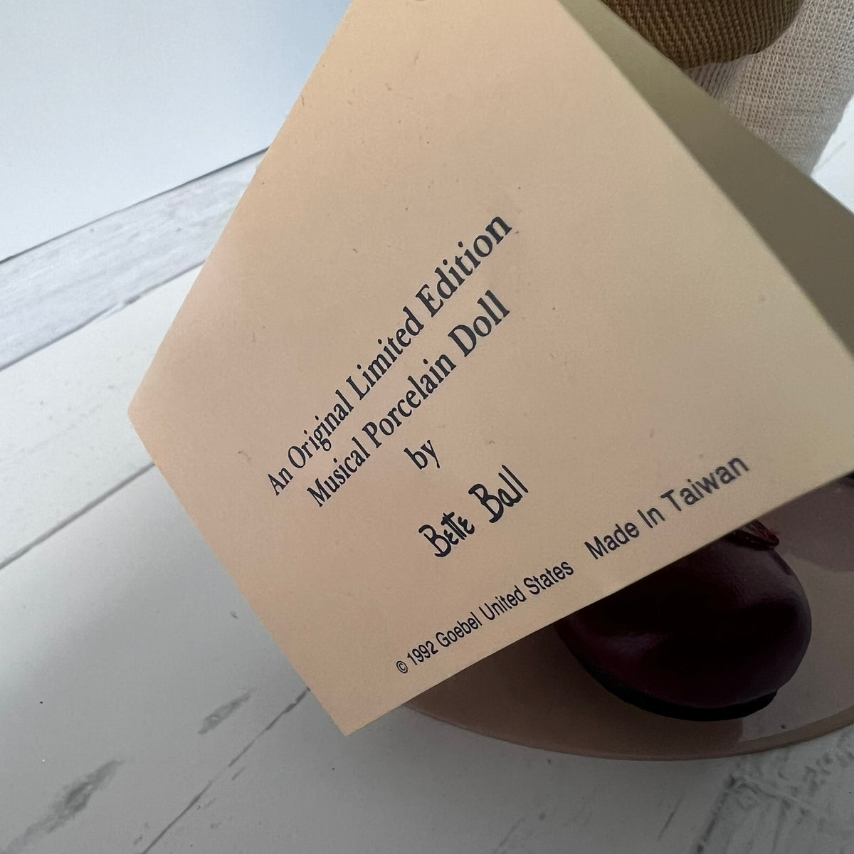 A close-up of a beige paper tag printed with 'An Original Limited Edition Musical Porcelain Doll by Bét Bell' and 'Made In Taiwan', resting over a dark purple porcelain doll on a white wooden surface.