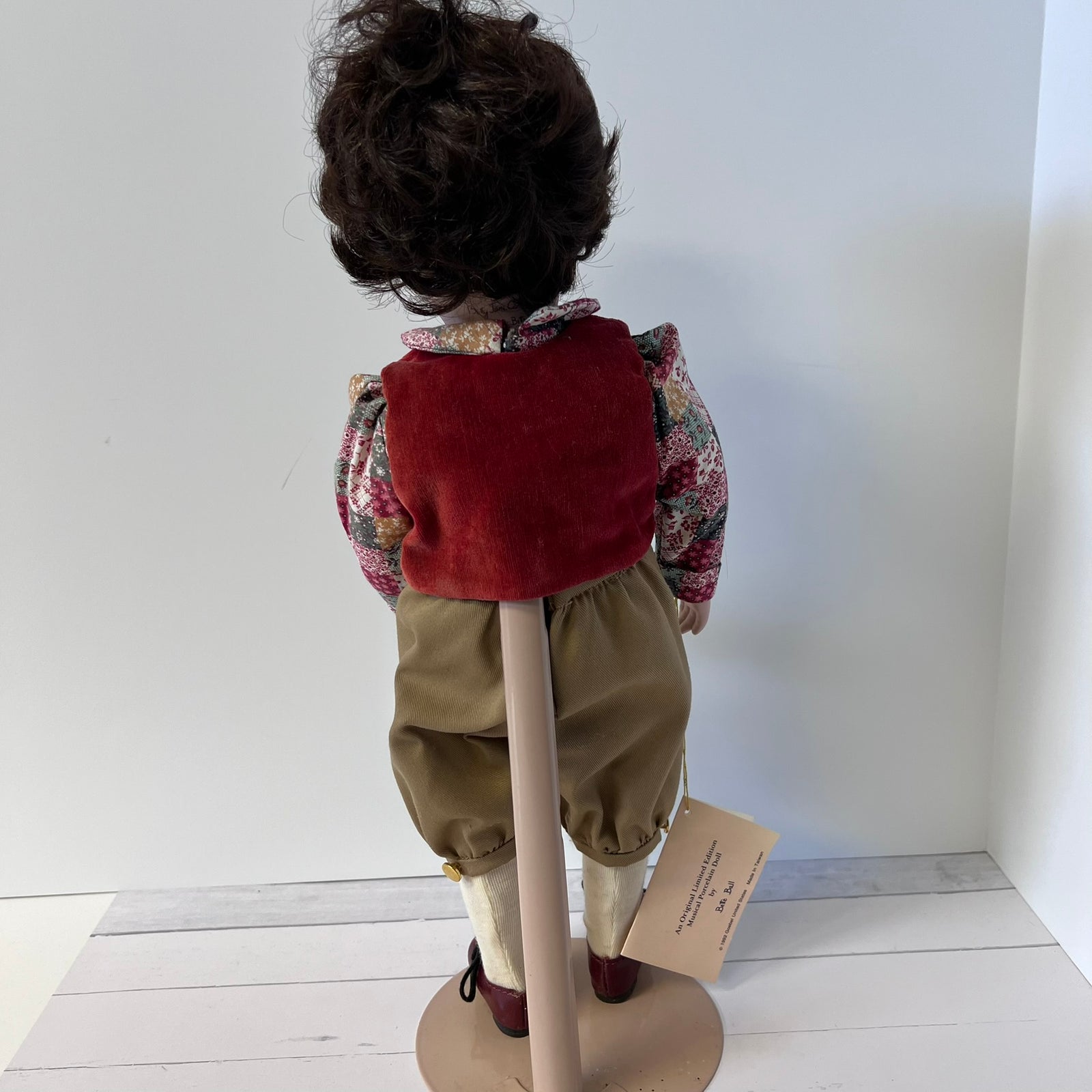 A vintage collectible doll viewed from behind, wearing a red velvet vest, a floral long-sleeve shirt, tan trousers, and dark shoes, mounted on a display stand.