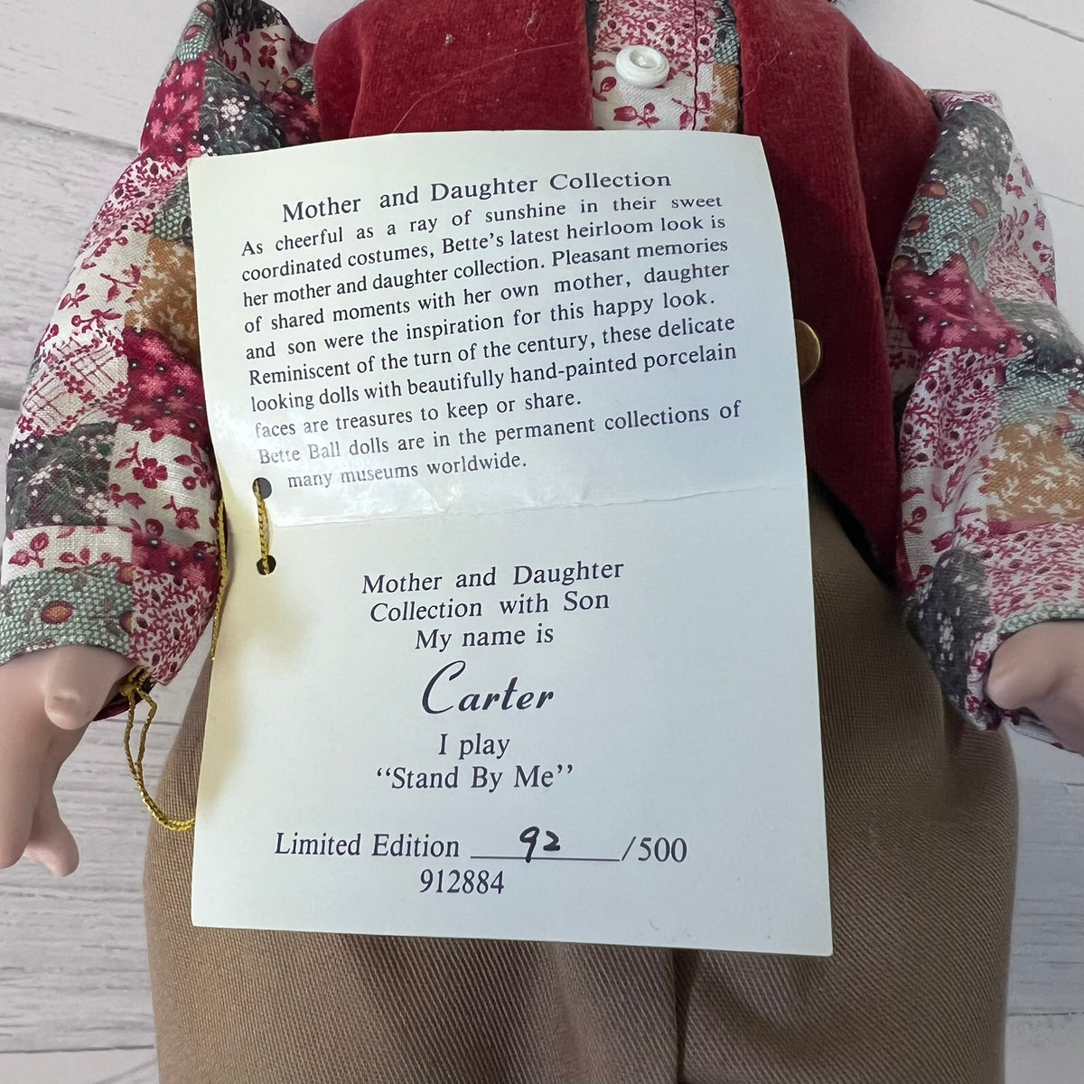A close-up of a doll with a cream-colored face and hands, wearing a red vest and a floral dress, holding or next to a printed paper tag that reads 'Mother and Daughter Collection' with the name Carter.