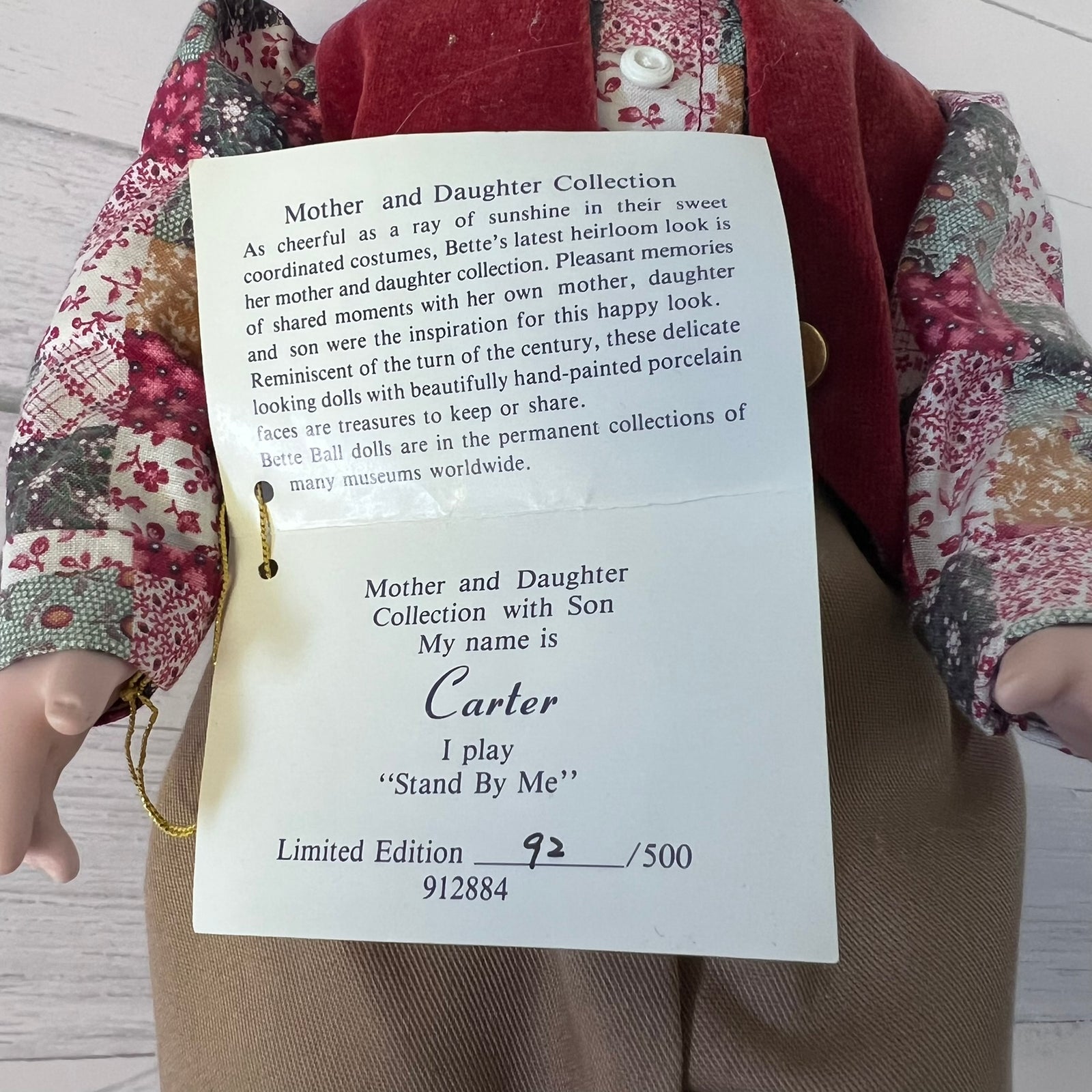 A close-up of a doll with a cream-colored face and hands, wearing a red vest and a floral dress, holding or next to a printed paper tag that reads 'Mother and Daughter Collection' with the name Carter.