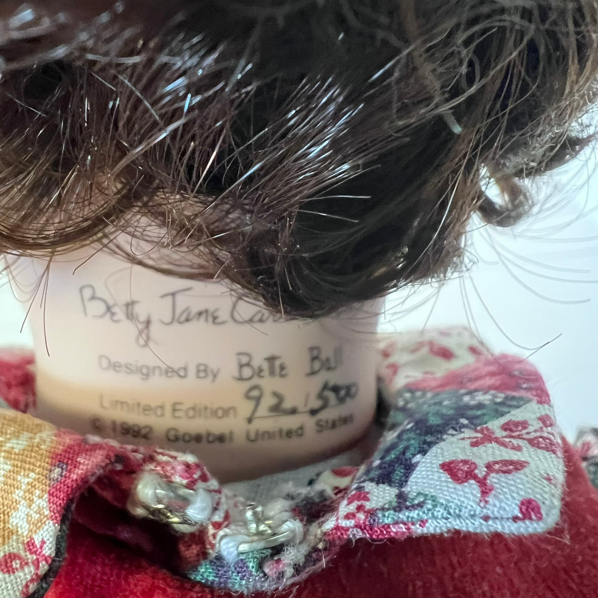 Close-up of a vintage Betty Jane doll showing its brown hair, cream-colored neck label with handwritten edition details, and a red floral patterned fabric collar.