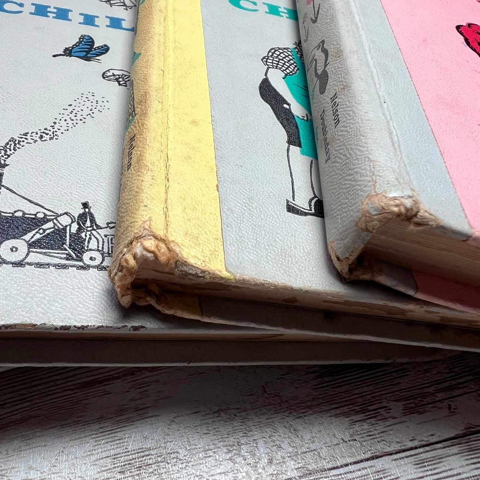 A close-up of vintage hardcover books stacked on a rustic wooden surface. The books show heavy wear and tear along the edges, with one featuring a yellow spine and others showing light blue and pink covers with illustrated characters.