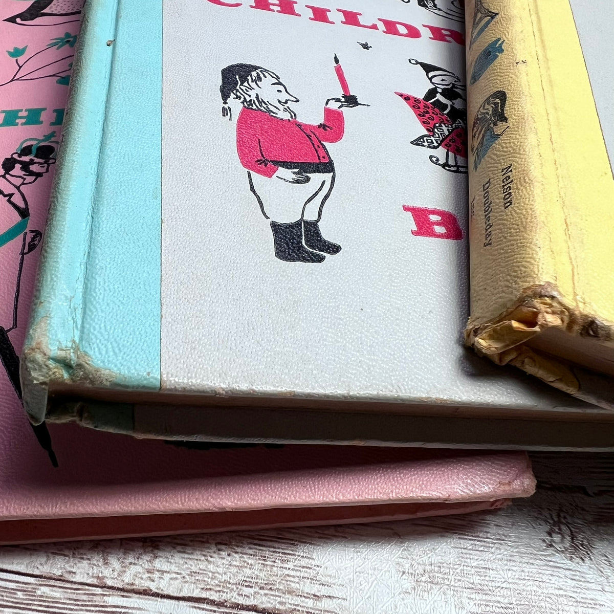 A close-up of a vintage children's book with a light blue spine and a cream cover featuring a black-and-red illustrated figure, stacked on other pink and yellow books on a whitewashed wood surface.