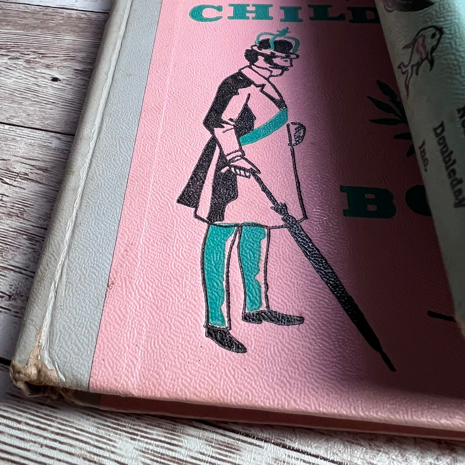 A vintage hardcover book or notebook with a pink cover featuring a black-and-white illustrated figure and teal accents, photographed on a rustic whitewashed wood surface.