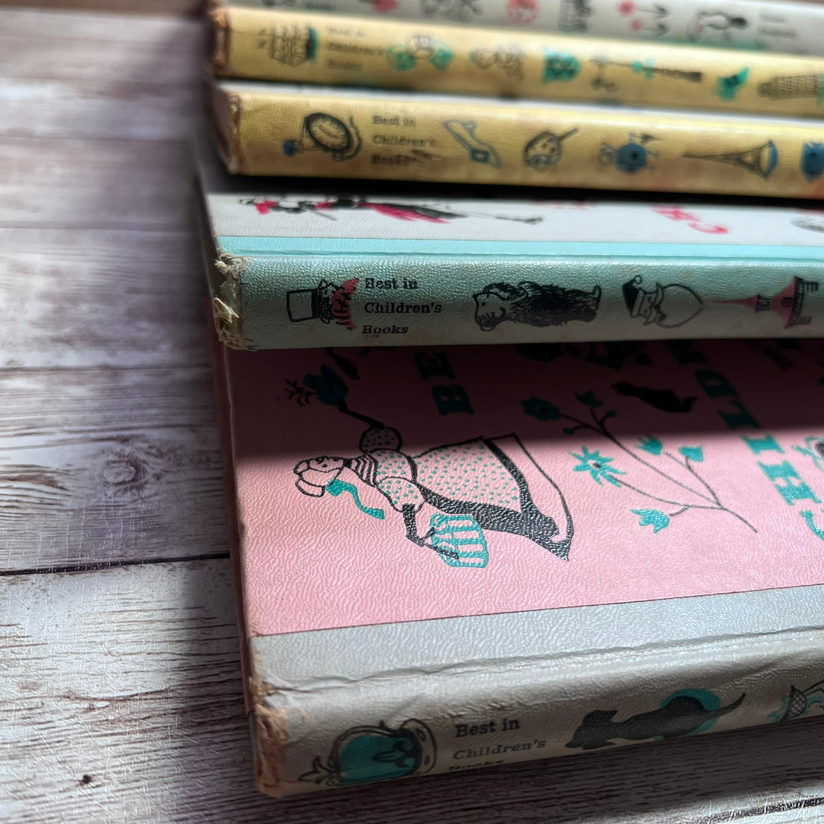 A stack of vintage children's books with illustrated spines and pastel covers, photographed on a rustic wooden surface.