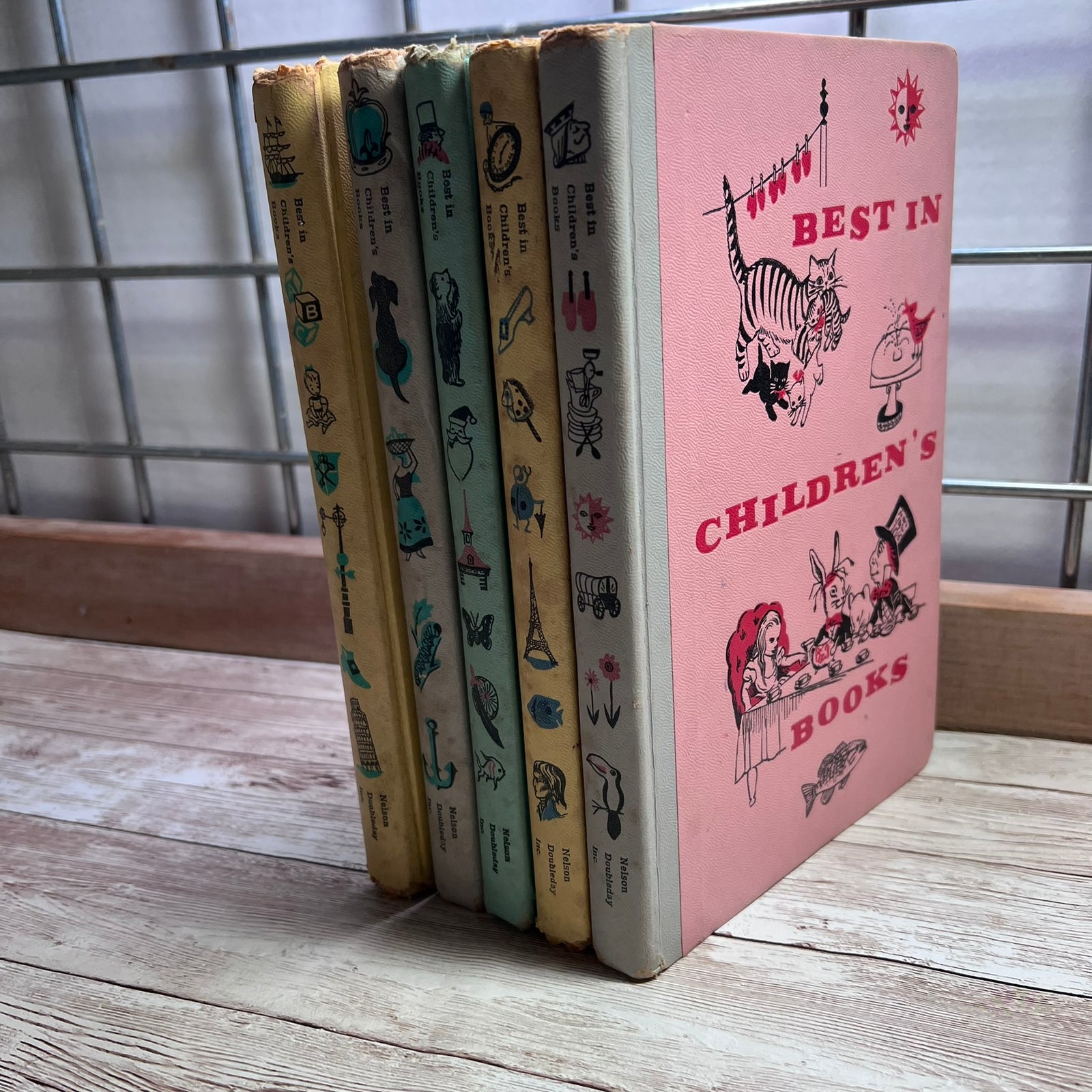 A set of vintage children's books standing upright on a wooden surface in front of a metal grid window. The front book has a pink cover with red text reading 'BEST IN CHILDREN'S BOOKS' and black-and-white illustrations, including a cat and a rabbit.