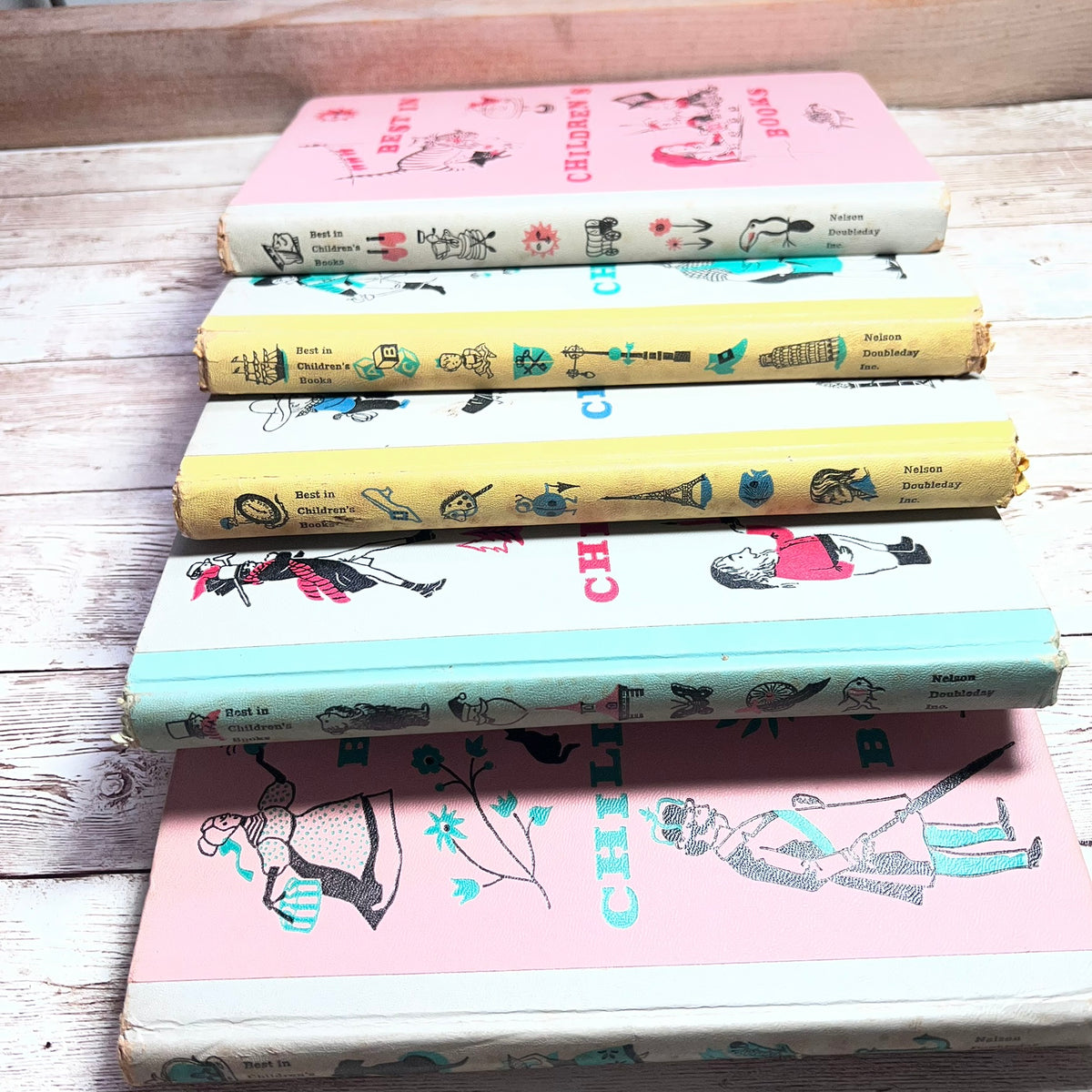A stack of five vintage children's books with illustrated spines, arranged on a rustic whitewashed wooden surface. The spines show pastel colors including pink, yellow, blue, and white, with decorative drawings and text such as 'Children's Book' and 'Childhood'.