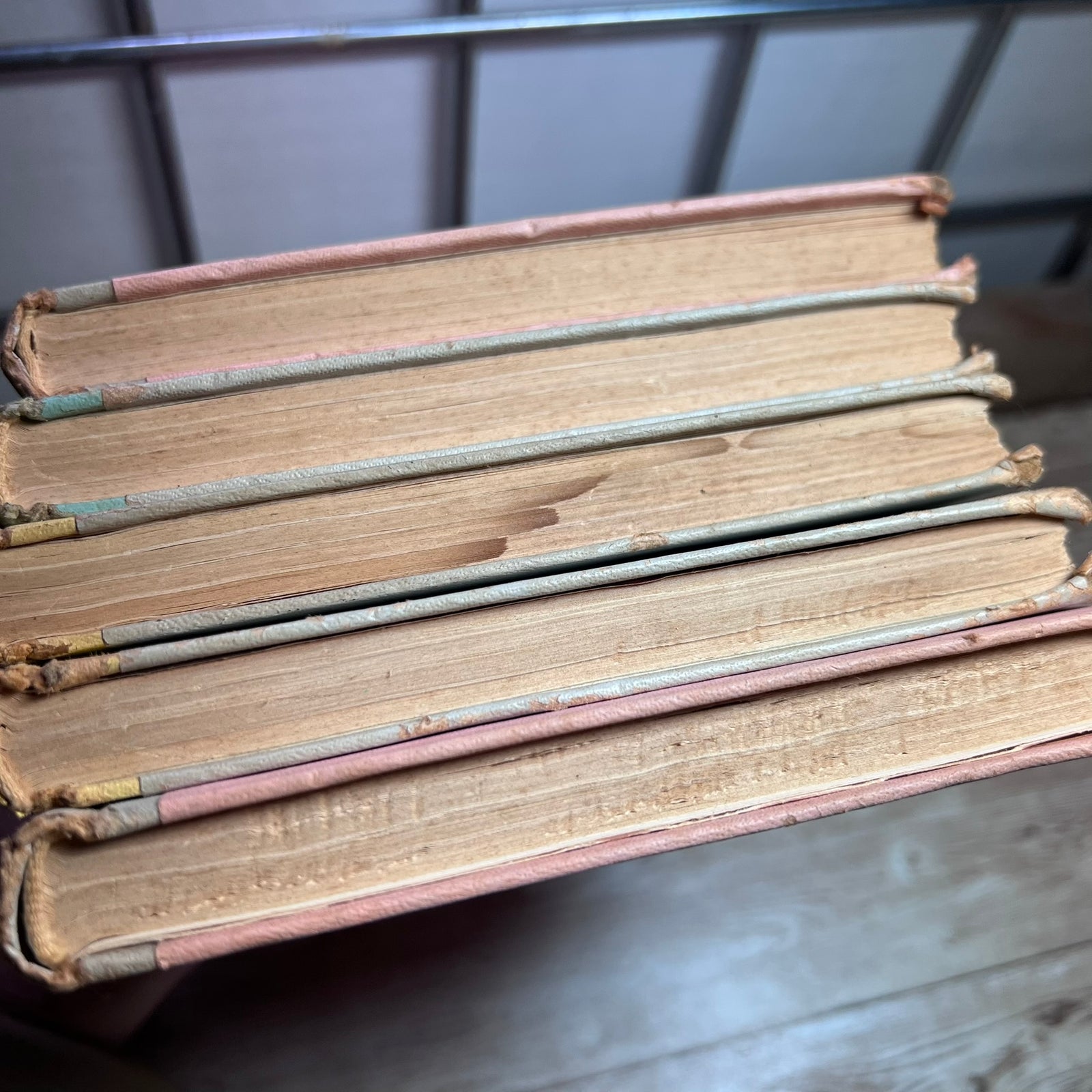 A close-up of a stack of vintage hardcover books with worn, tan pages and pinkish-red spines, resting on a wooden surface.