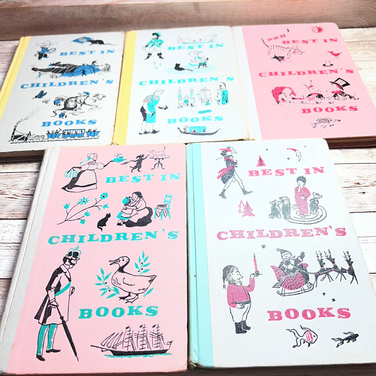 Five vintage hardcover children's books arranged on a light wood surface. The covers are primarily pink, cream, and light blue, featuring whimsical illustrations and the text 'BEST IN CHILDREN'S BOOKS'.