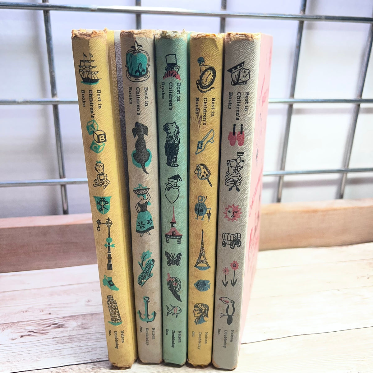A set of five vintage children's books standing upright on a light wooden surface in front of a metal grid window. The spines show illustrated covers with titles including 'The Sea Captain', 'The Sea Witch', 'The Sea Monster', 'The Sea Princess', and 'The Sea Flower'.