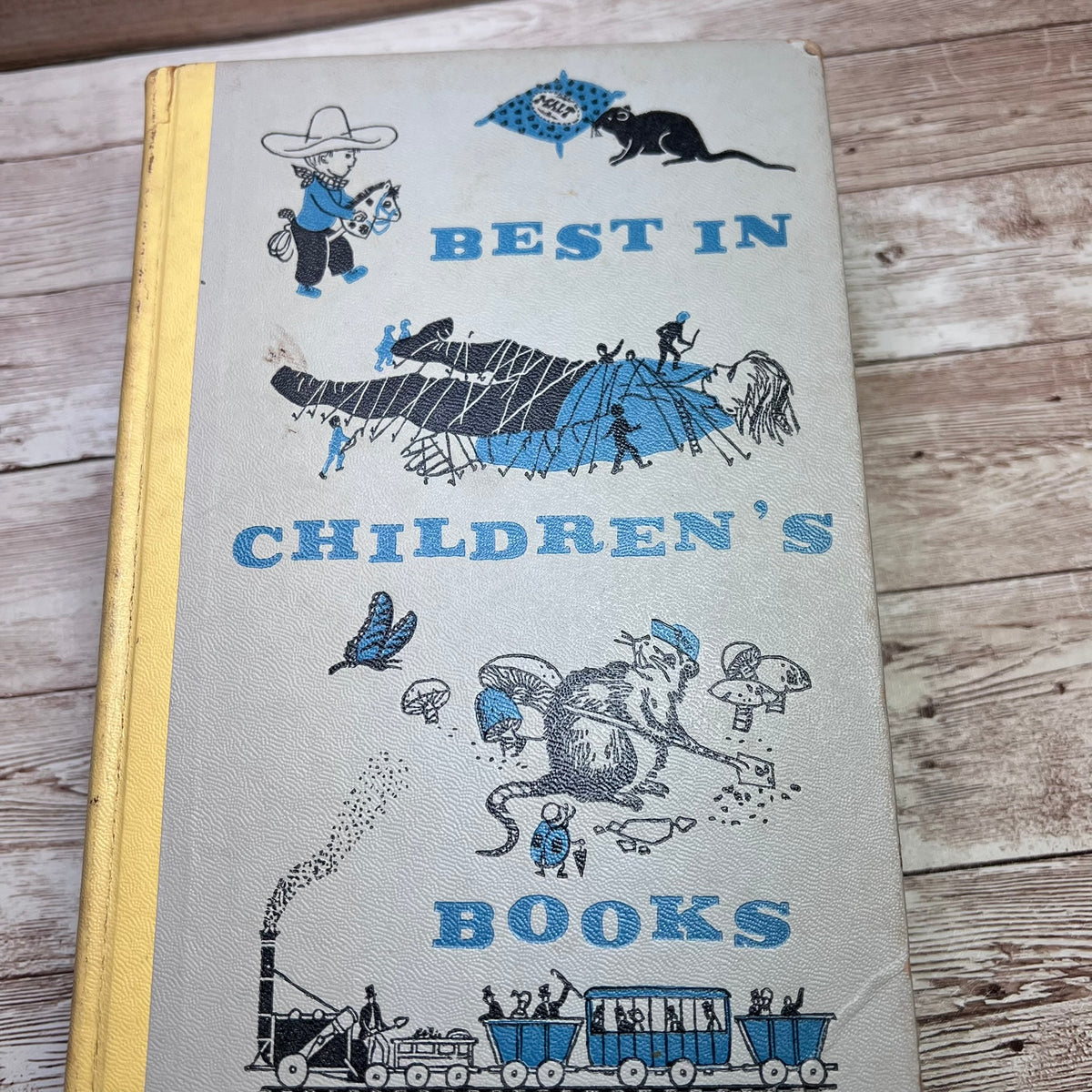 Vintage hardcover children's book titled "BEST IN CHILDREN'S BOOKS" with blue illustrations and a yellow spine, photographed on a rustic wood surface.