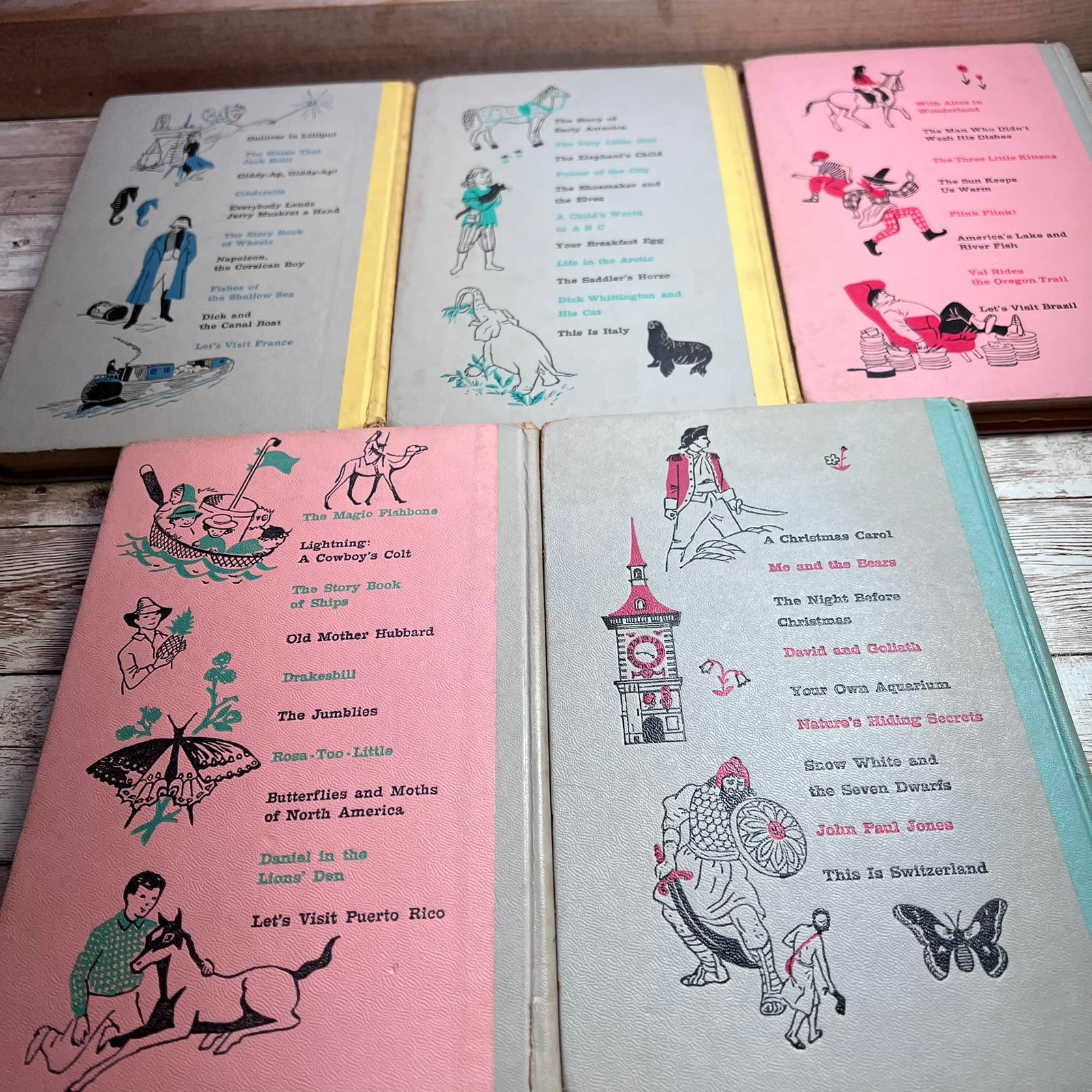 Five vintage children's activity books with illustrated covers are arranged on a light wood surface. The covers are in pastel pink, gray, and blue-green tones with black-and-white and colored illustrations and lists of activities and reading stories.