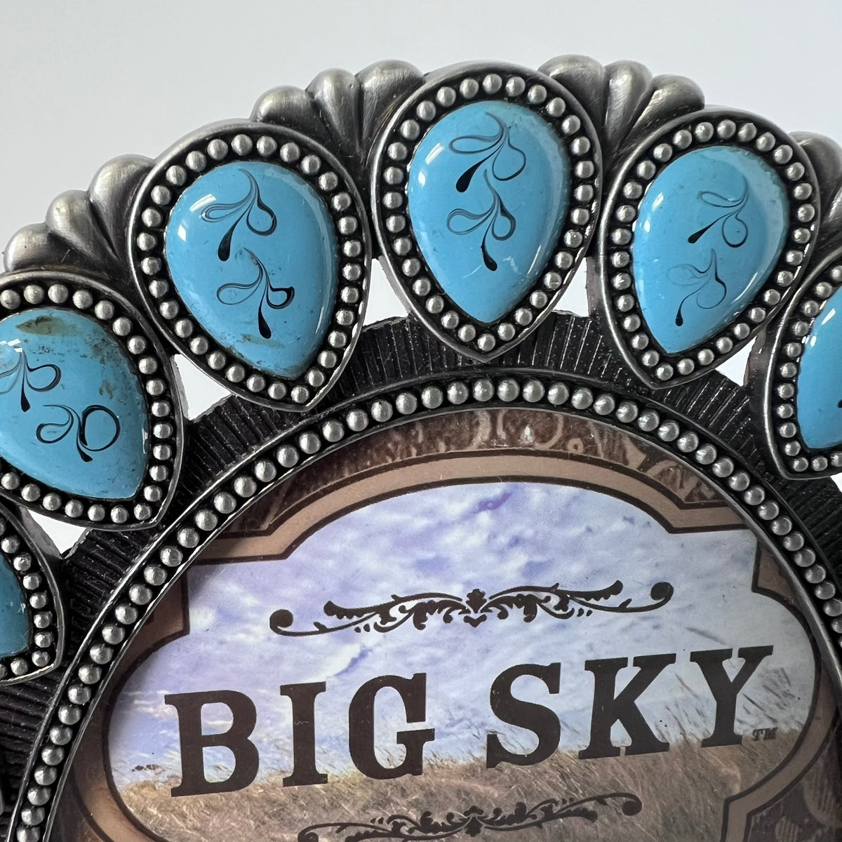 A close-up of a decorative oval picture frame with silver-toned metal detailing, turquoise blue inlay stones, and a central printed insert reading “BIG SKY.”.