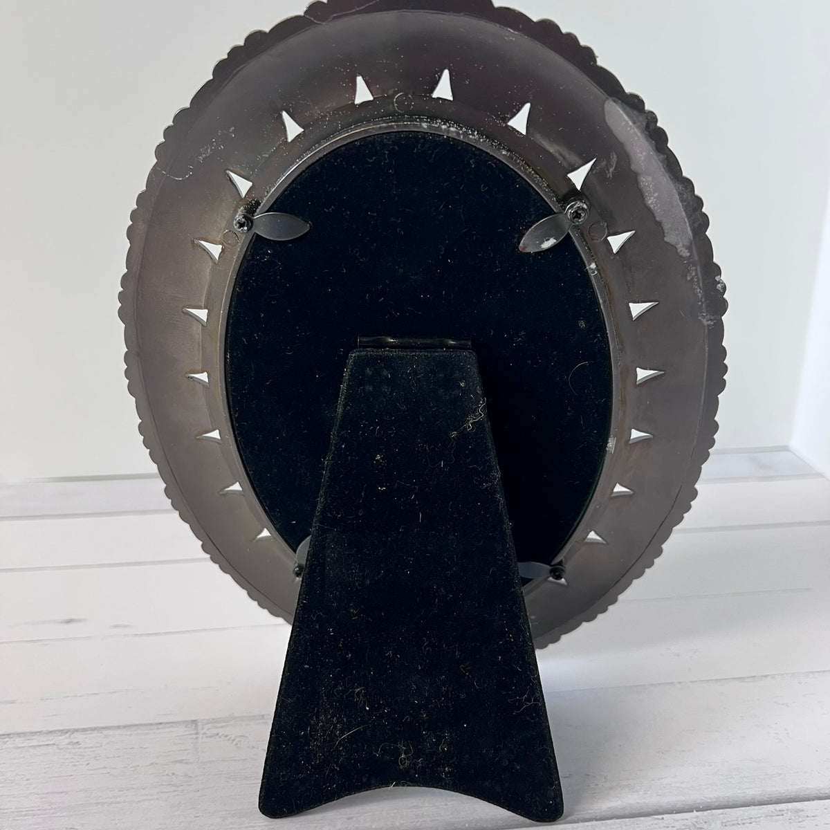 A black metal picture frame with a scalloped outer edge and a black velvet display stand, shown from the back on a white wooden surface.