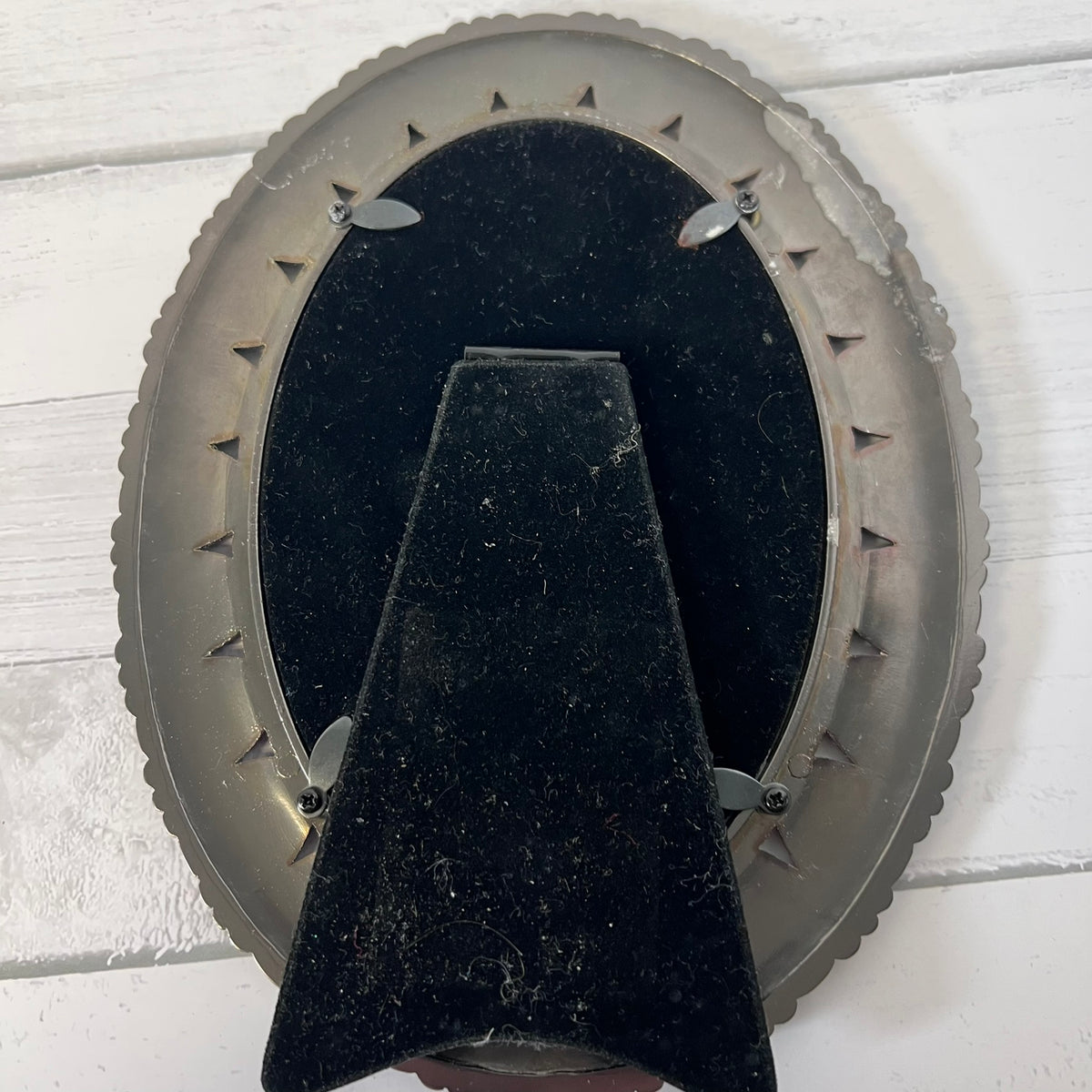 The back of an oval wall-mounted picture frame, showing a black velvet backing and a central support bracket, mounted on a white painted wood plank wall.