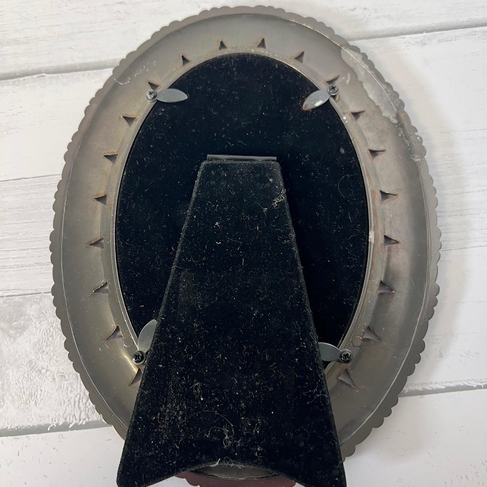 The back of an oval wall-mounted picture frame, showing a black velvet backing and a central support bracket, mounted on a white painted wood plank wall.