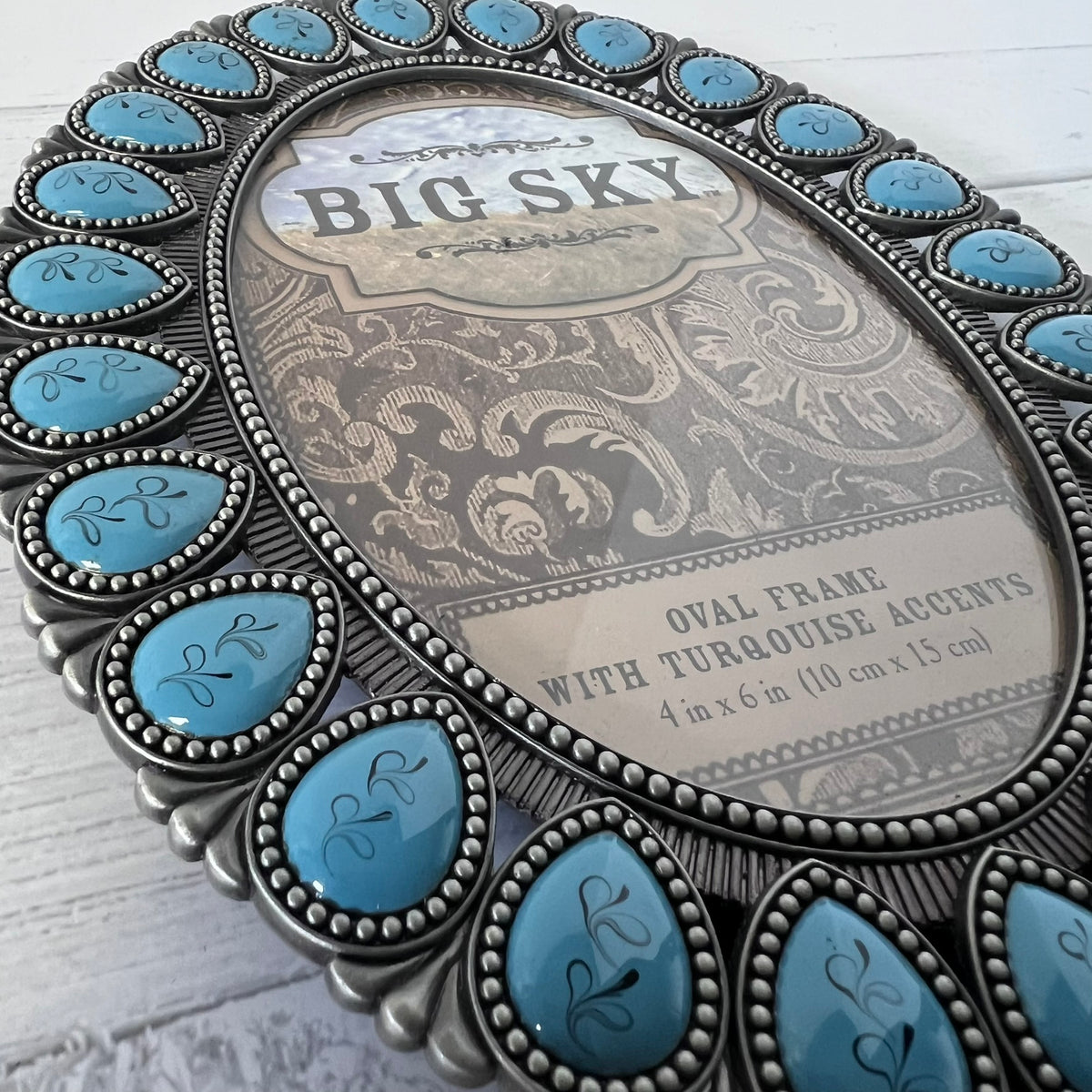An oval picture frame with a distressed silver-toned metal border and turquoise stone accents, shown on a white wood background. The center insert reads 'BIG SKY'.