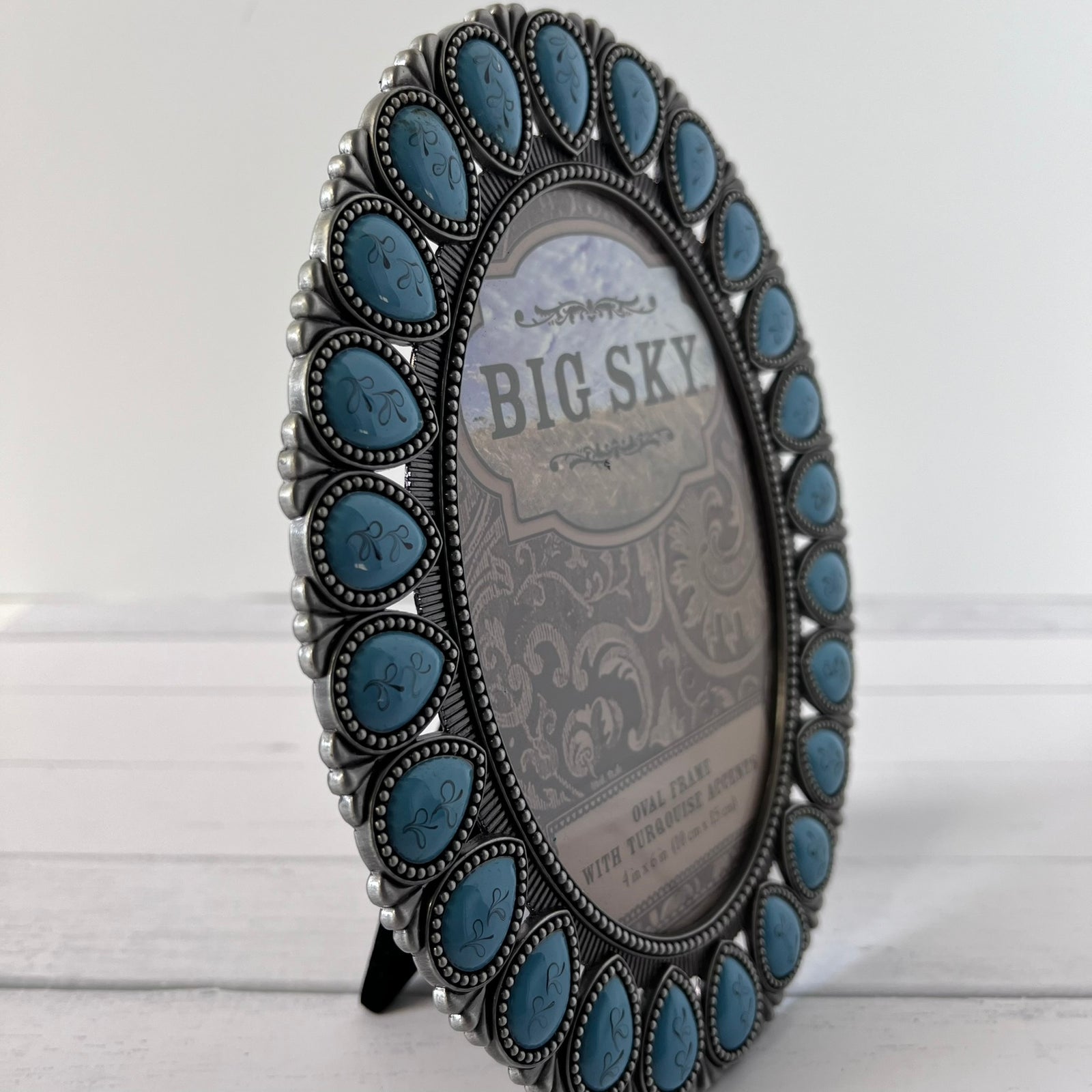 An oval silver picture frame decorated with a border of turquoise-blue stone inlays, standing on a small black easel against a white wood background.