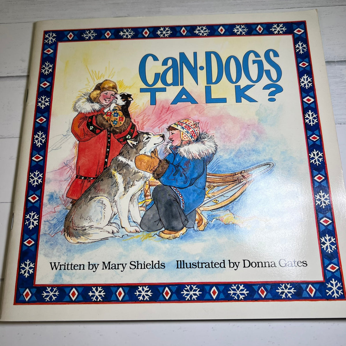 Children's book titled "Can Dogs Talk?" with a cream cover, blue decorative border, and an illustration of a child in a blue coat kneeling with a dog while another person in a red coat holds a black dog. Text at the bottom reads "Written by Mary Shields Illustrated by Donna Gates.