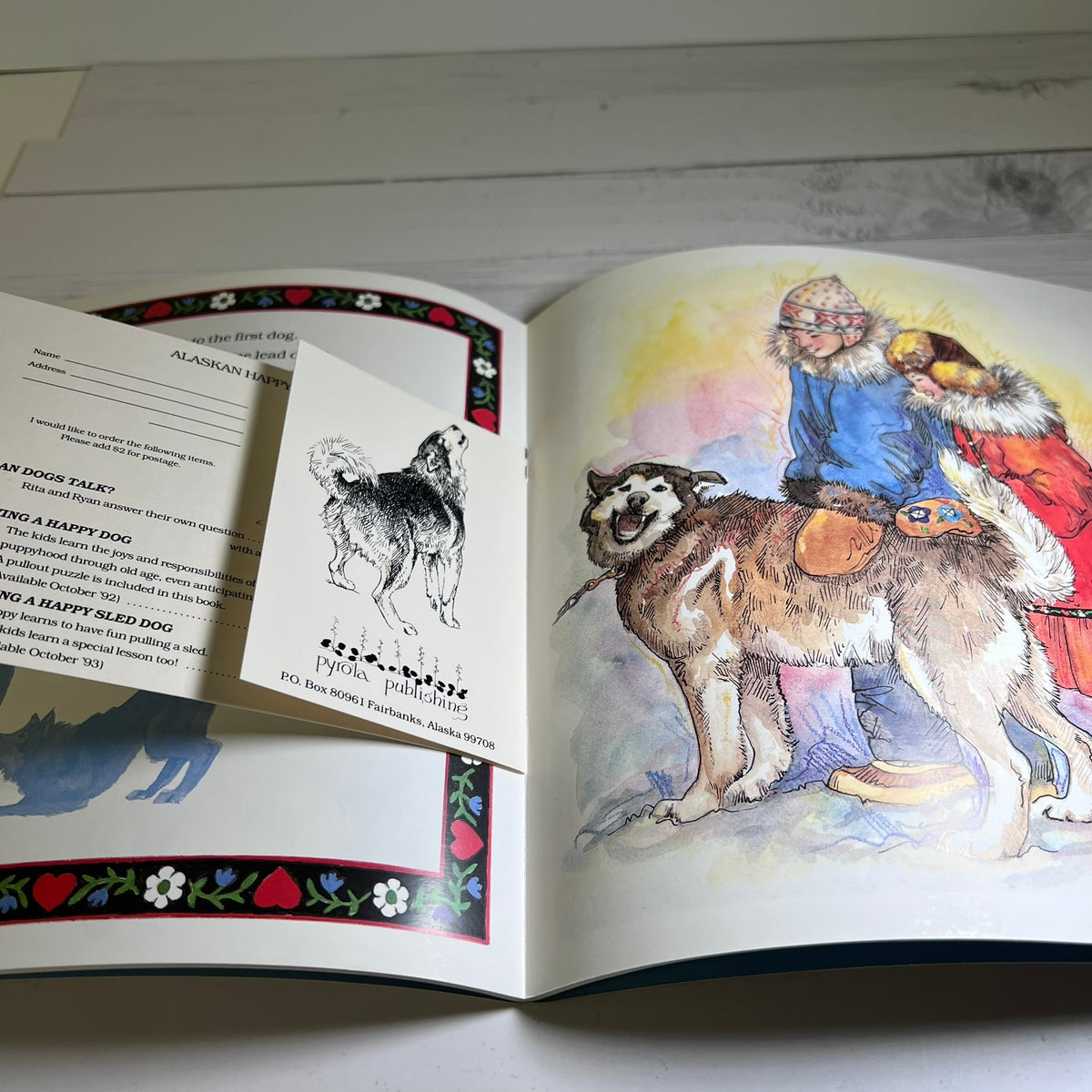 An open children's book or booklet laid on a light surface, showing printed text on the left page and a watercolor illustration on the right page of a dog wearing a winter outfit with a red coat and blue jacket.