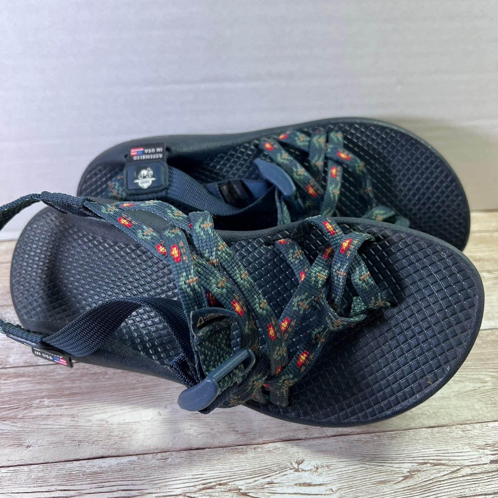 A pair of black Perrier outdoor sandals with camouflage-patterned woven straps and small red accents, resting on a light wood surface.