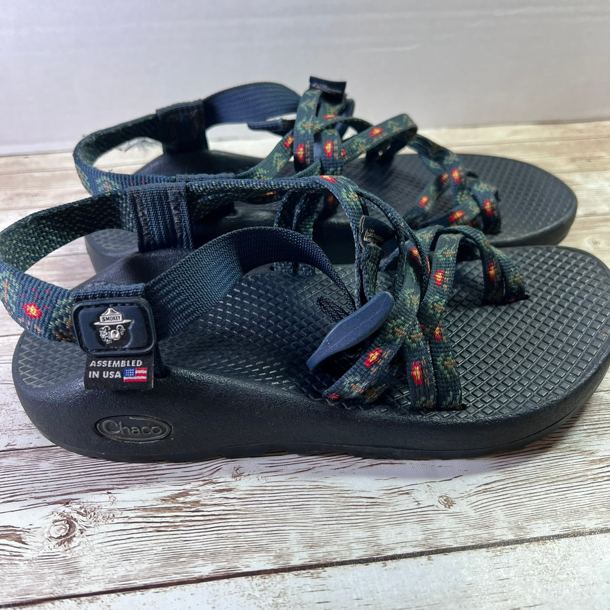 A pair of Chaco sandals with dark navy straps and footbeds, featuring a woven pattern on the front straps with small red and orange accents. The sandals are placed on a light-colored wooden surface.
