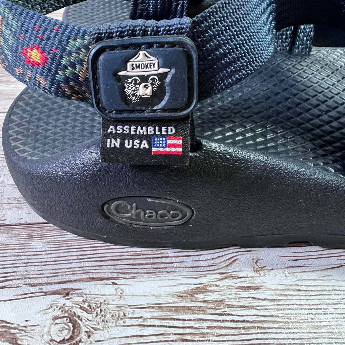 A close-up of a black Chaco sandal with a patterned blue and red woven strap, featuring a black plastic buckle with a Smokey bear logo and an 'Assembled in USA' tag, resting on a rustic wooden surface.