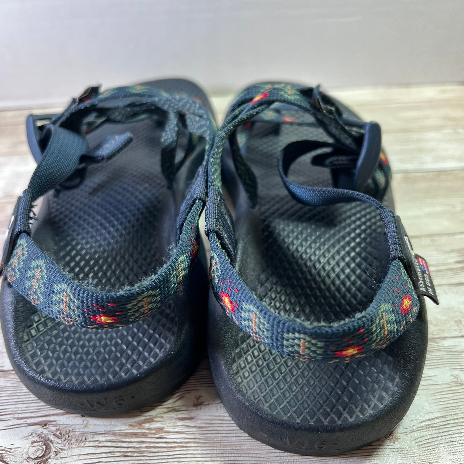 A pair of Teva sandals with dark gray/black textured footbeds and patterned straps featuring blue, green, and orange accents, resting on a light wood surface.