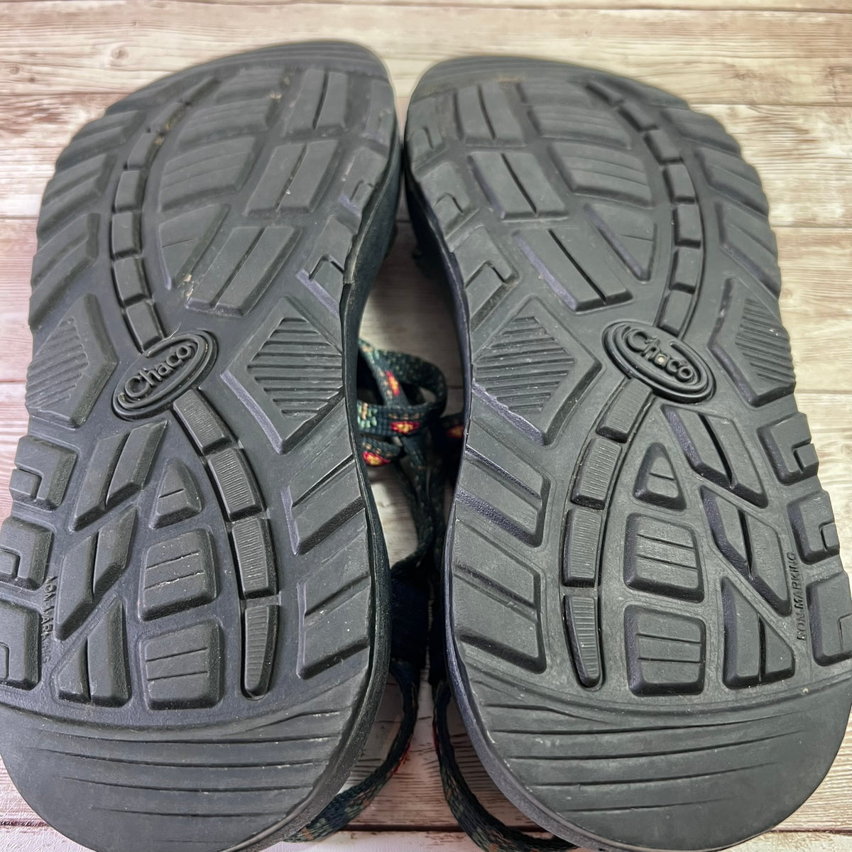 A pair of black Chaco sandals shown from the bottom, displaying the treaded soles on a light-colored wooden floor. The Chaco logo is clearly visible on the center of each sole.