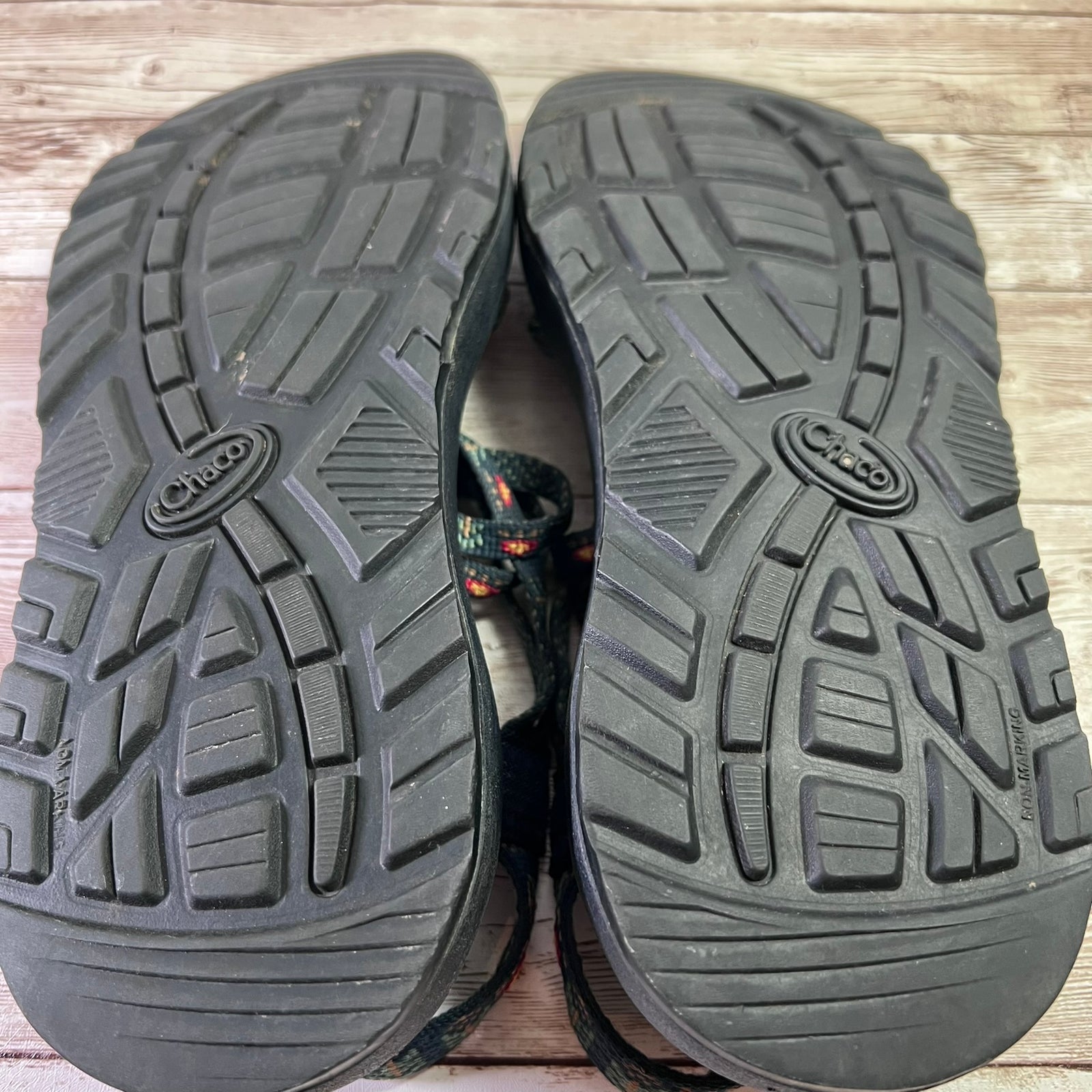 A pair of black Chaco sandals shown from the bottom, displaying the treaded soles on a light-colored wooden floor. The Chaco logo is clearly visible on the center of each sole.
