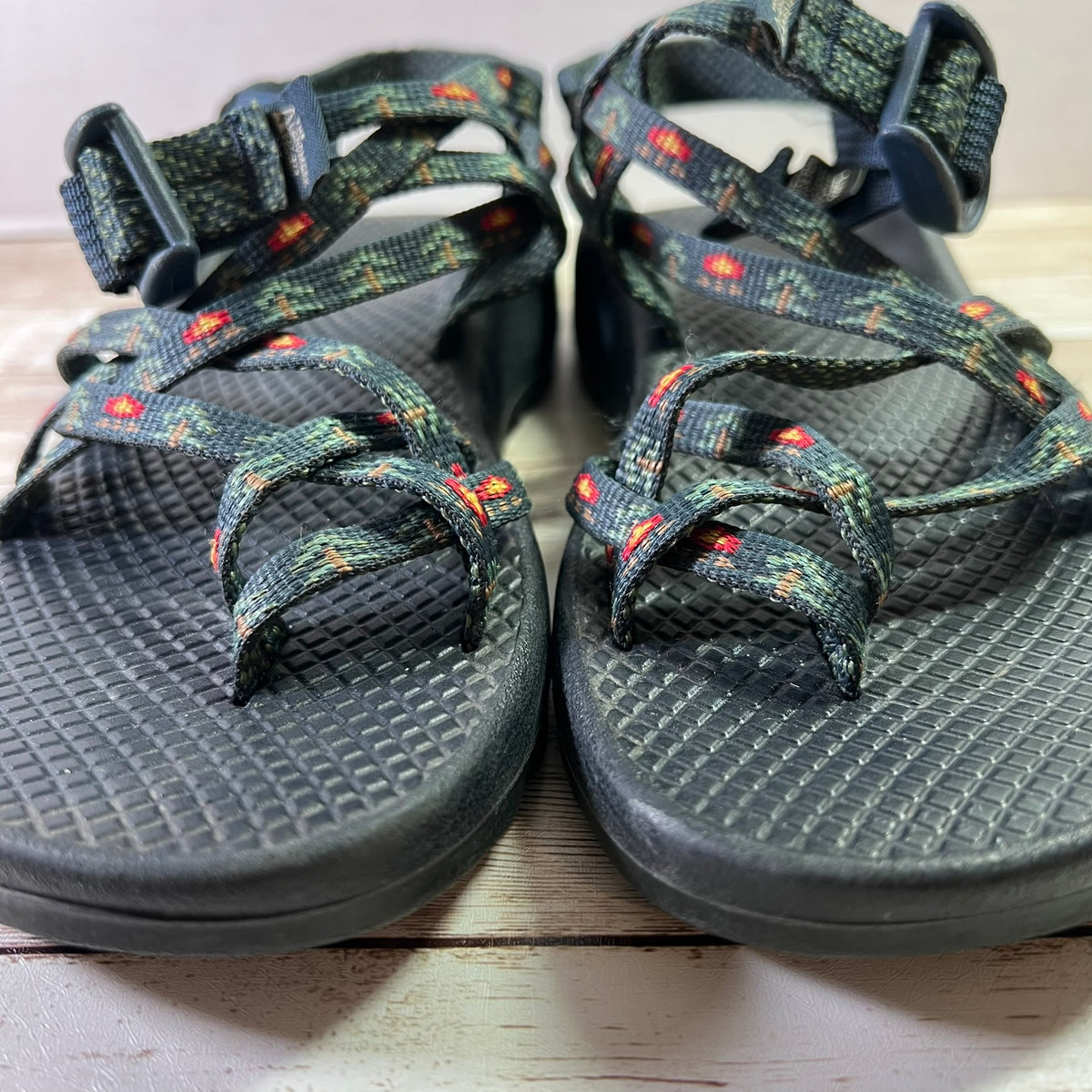 A pair of Chaco-style athletic sandals with dark gray textured footbeds and woven straps featuring a green, black, and red pattern, resting on a light wood surface.