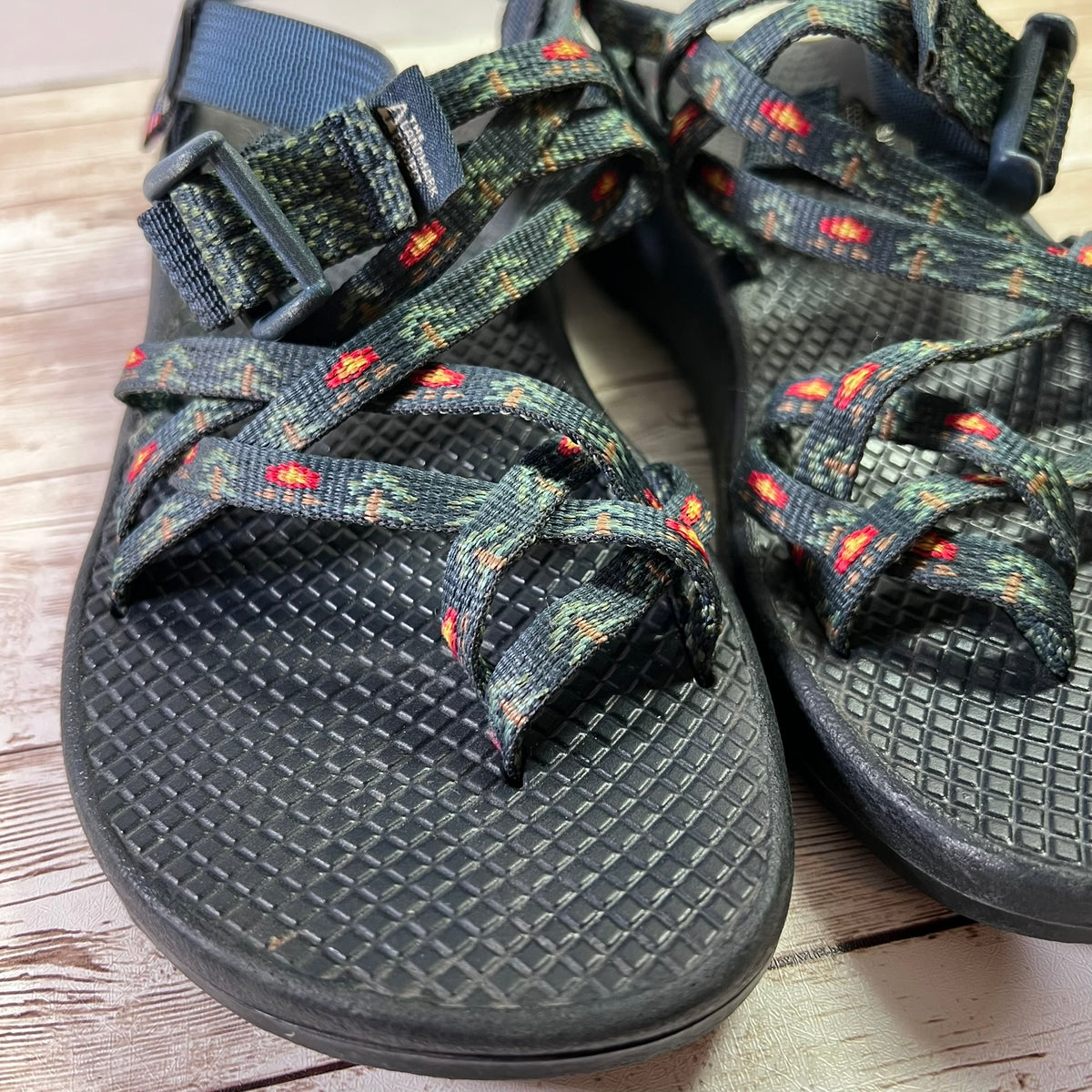 A pair of black woven athletic sandals with colorful patterned straps and textured black footbeds, resting on a light wood surface.