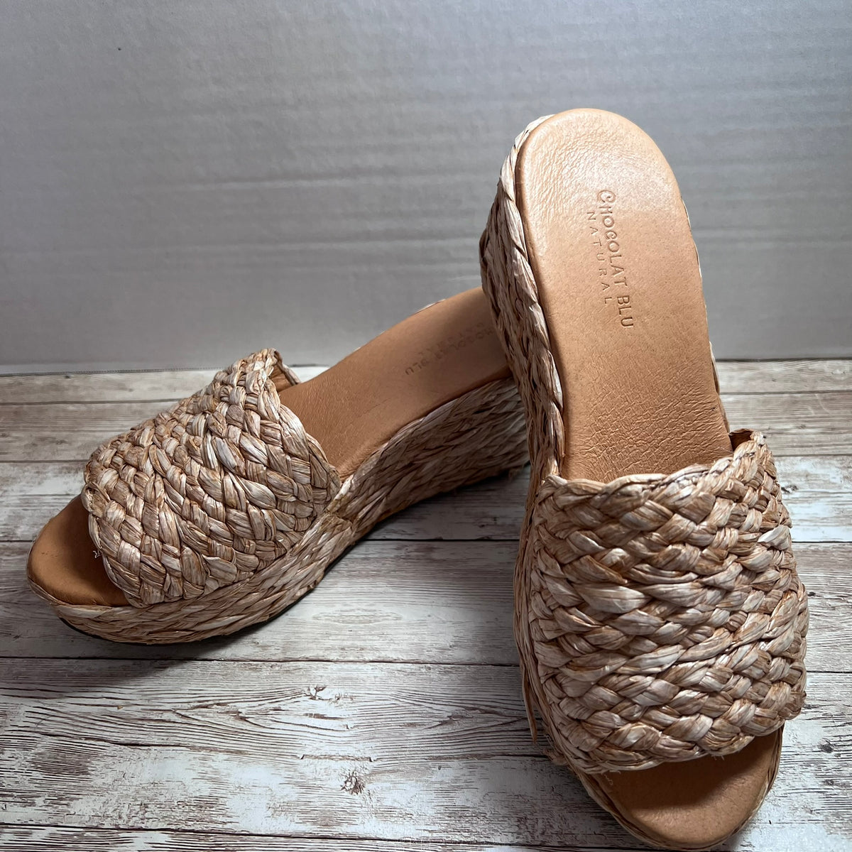 A pair of women's slide sandals with woven straw-like upper straps and wedge platforms, shown on a rustic wood surface against a light gray wall. The insole is tan and shows the brand name 'Goddard's Bay'.