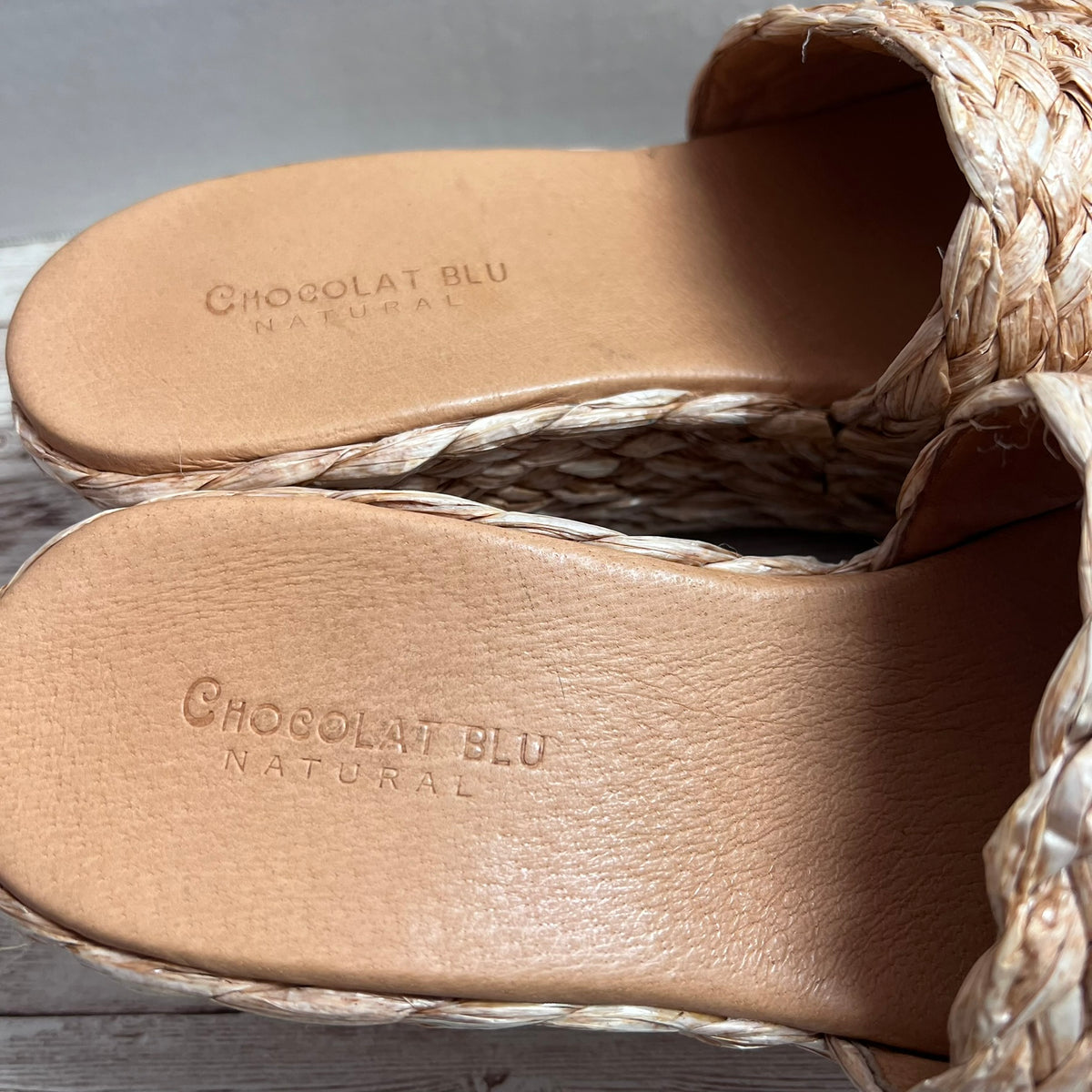 Close-up of the insoles of a pair of woven slide sandals, showing the embossed brand name 'Chocolat Blu Natural' on natural tan leather. The sandals feature braided jute straps and a matching braided outsole.