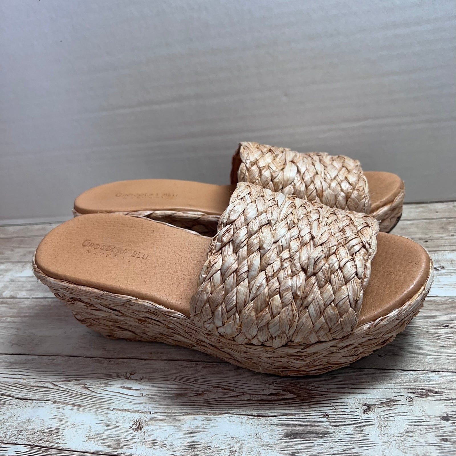 A pair of women's wedge slide sandals with woven straw straps and a braided jute platform sole, shown on a rustic wood floor against a gray wall.