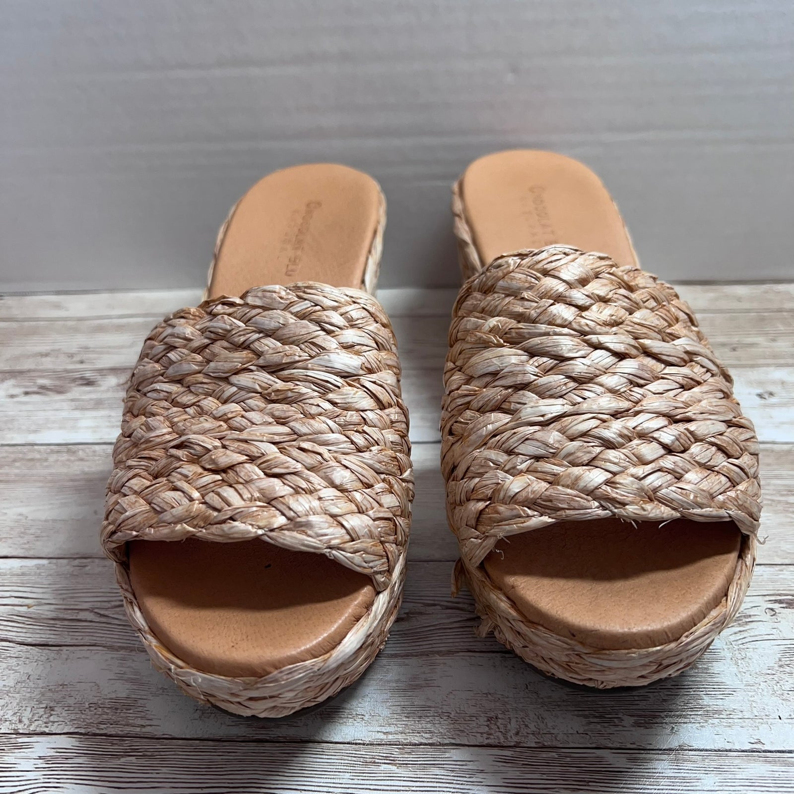A pair of woven straw slide sandals with platform soles, displayed on a light gray wood plank surface against a neutral gray background.