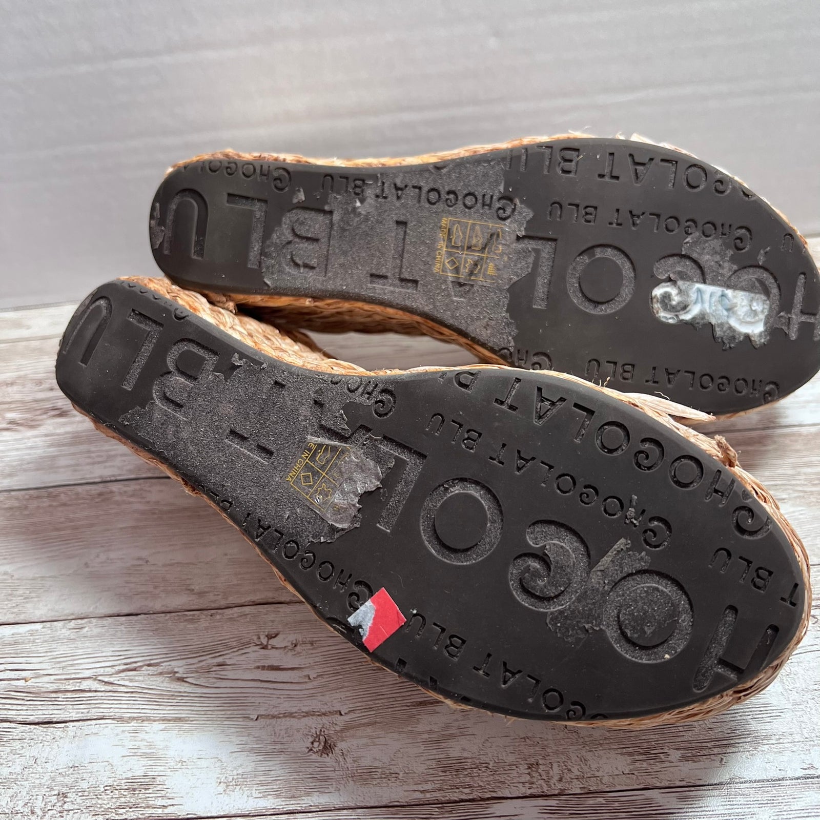 A pair of espadrille-style flat shoes shown from the sole side, resting on a light wood surface. The black rubber soles are embossed with the brand name 'Chocolat Bleu' repeatedly, and the natural jute-wrapped midsoles are visible along the edges.