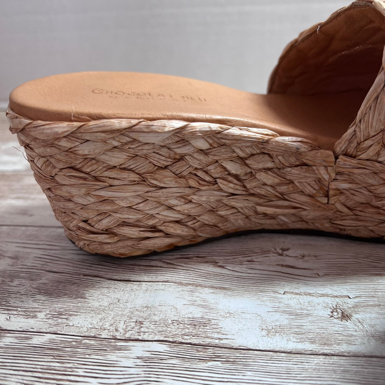 A close-up of a woven straw wedge sandal with a tan leather footbed, resting on a rustic white-washed wooden surface.