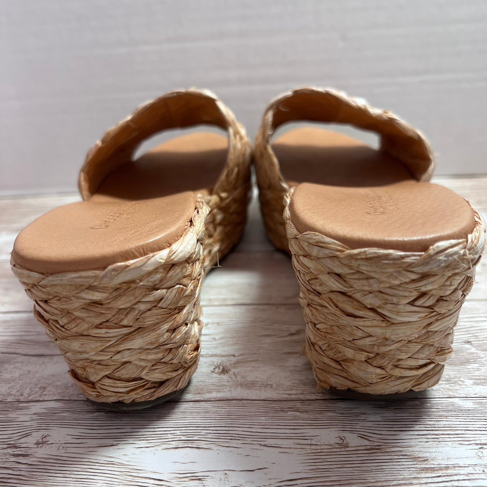 A pair of woven straw wedge sandals with tan leather straps and soles, shown from the back on a light wood surface against a white wall.